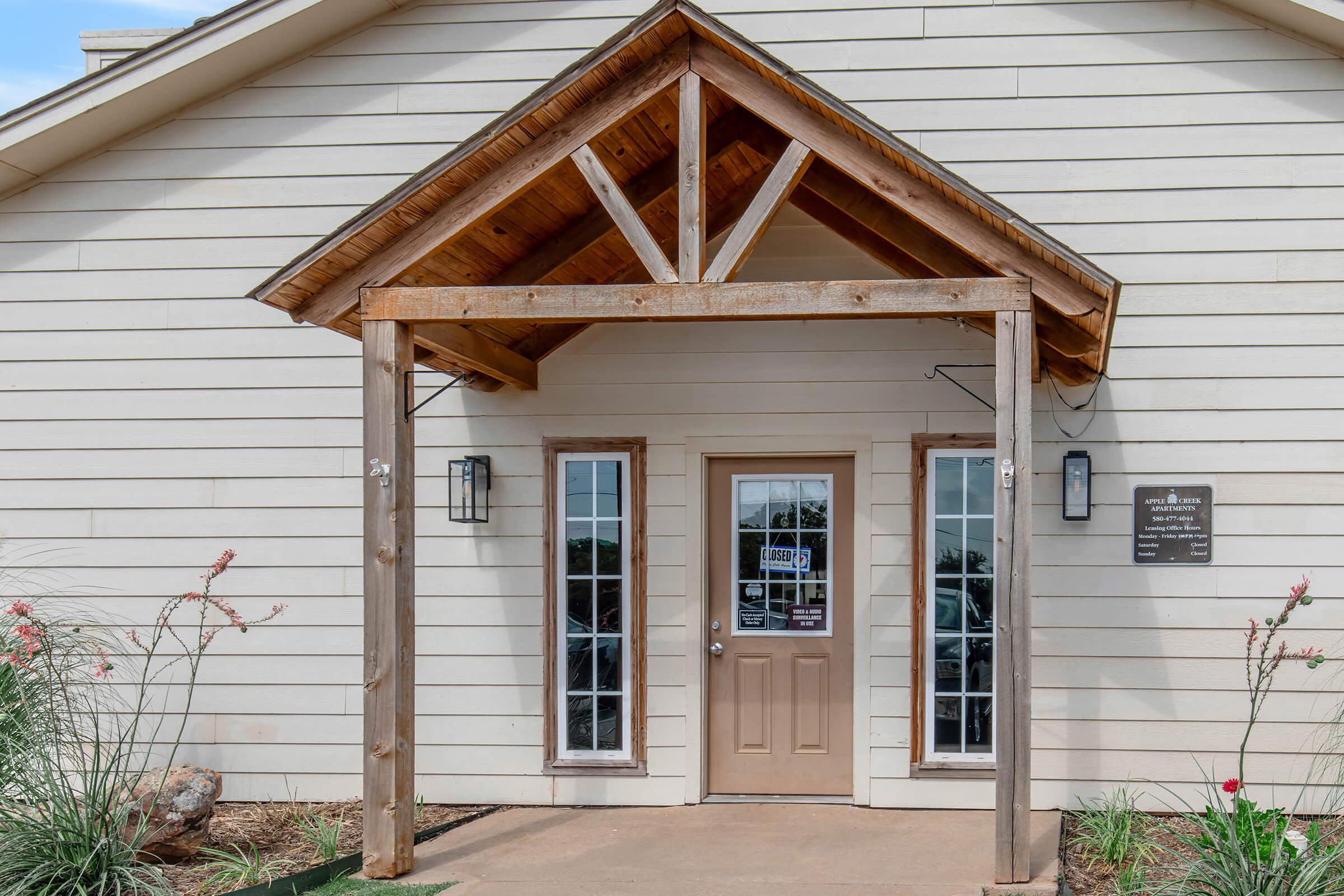 Entrance of a modern building featuring a wooden awning supported by large posts. The entrance has a single door with glass panels on either side and two light fixtures, along with a sign mounted next to the door. Surrounding the entrance are plants and decorative stones.