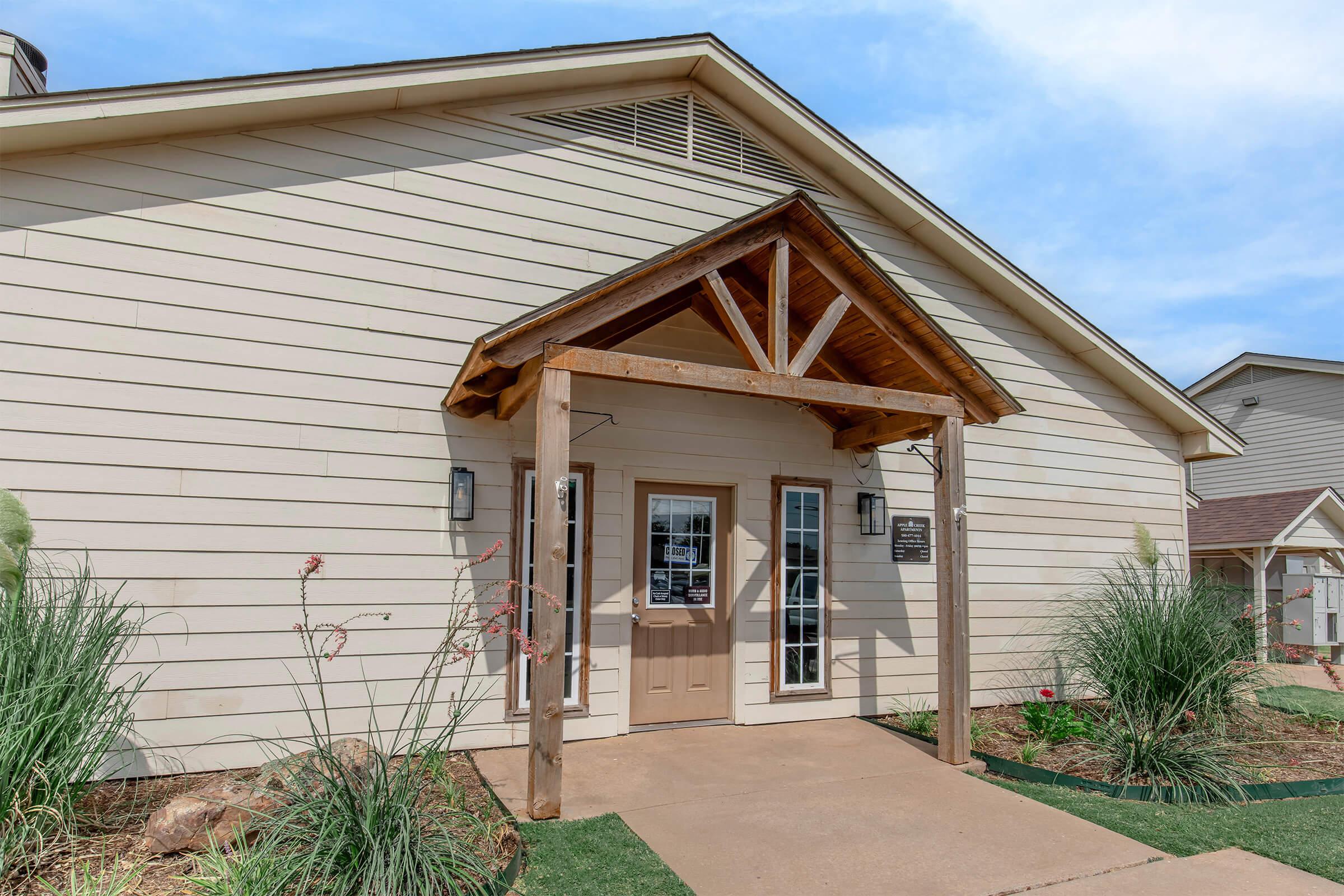 A light-colored building with a wooden porch featuring a triangular roof support. The entrance has double glass doors and two lantern-style lights on either side. Tall grass and plants line the pathway leading to the door. The sky is partly cloudy.