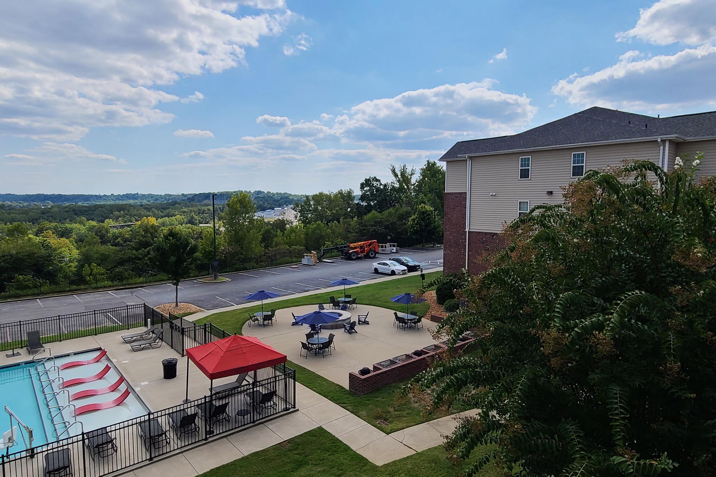 A view of an apartment complex with a swimming pool surrounded by lounge chairs and shaded tables. In the background, a landscaped area slopes down towards a river or lake, with trees lining the horizon and a cloudy sky overhead. A construction vehicle is parked in the parking lot.