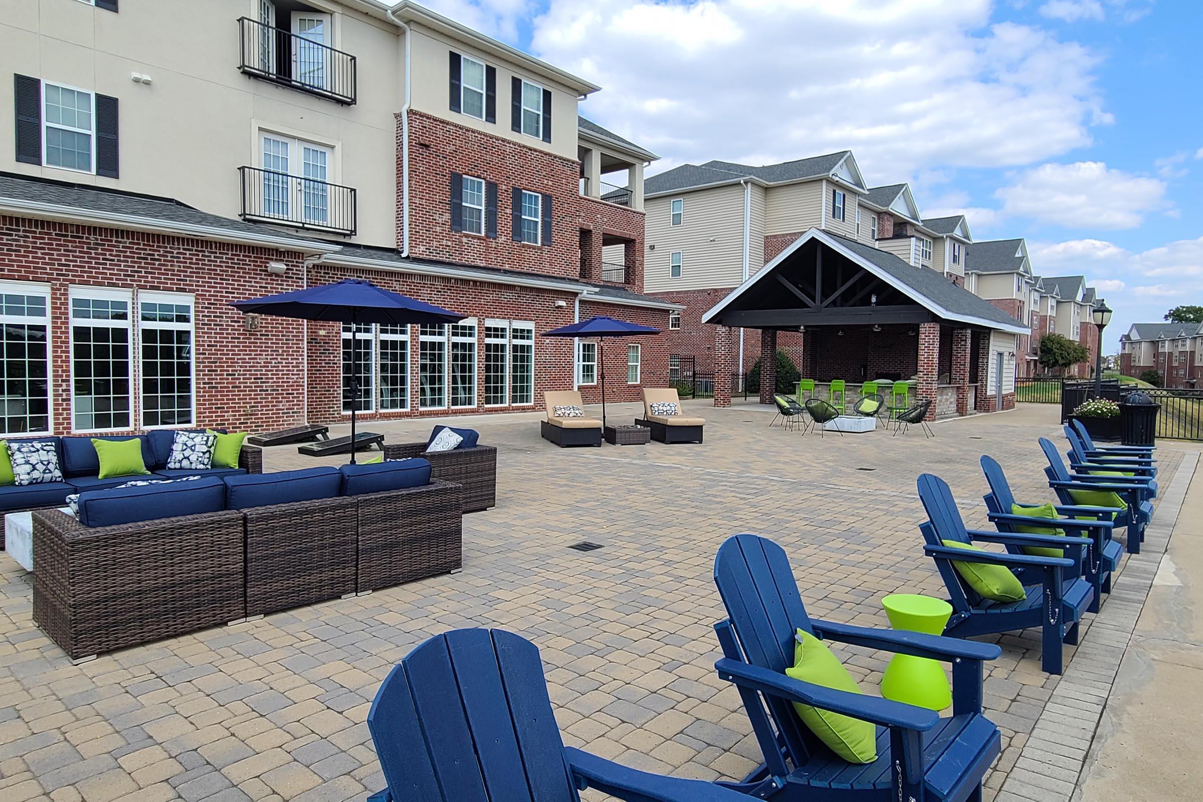 Outdoor lounge area featuring comfortable seating with blue cushions and green accents, surrounded by brick buildings. A covered pavilion is visible in the background, equipped with additional seating. The space is designed for relaxation and social gatherings, set against a clear blue sky.