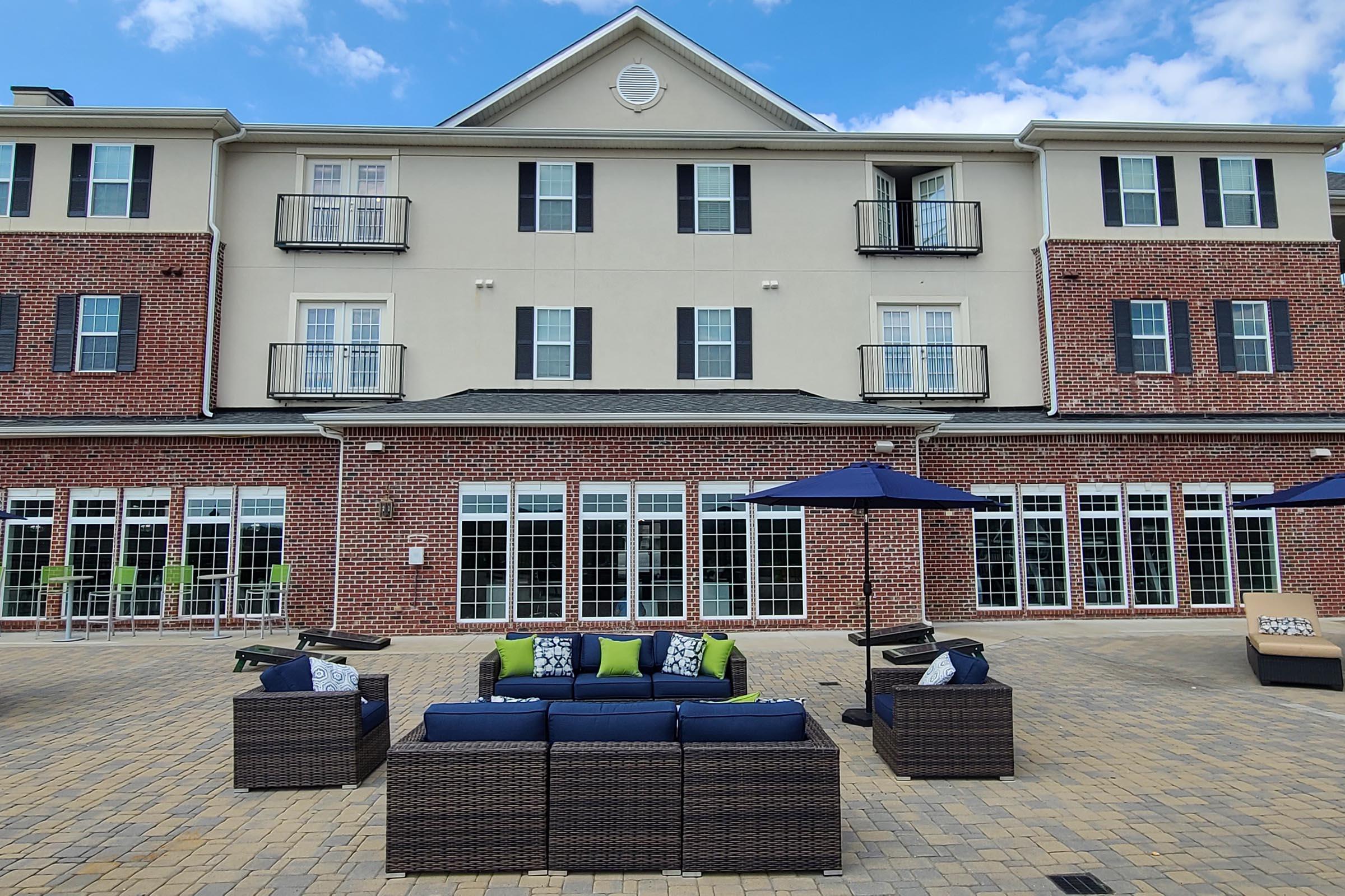 An outdoor patio area featuring a seating arrangement with dark wicker furniture and colorful cushions, surrounded by umbrellas. The backdrop includes a multi-story building with a brick facade and multiple windows, under a blue sky with a few clouds.