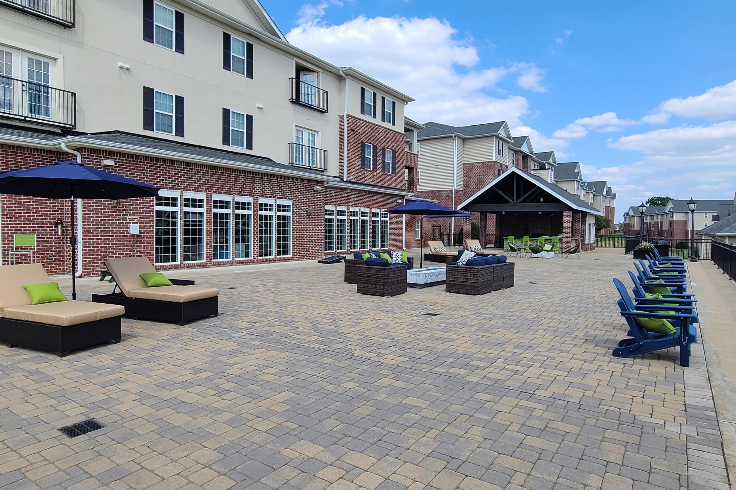 A spacious outdoor patio area featuring lounge chairs with green cushions, umbrellas for shade, and conversation seating near fire pits. The brick building in the background has large windows and a covered seating area, with a blue sky and fluffy clouds overhead.