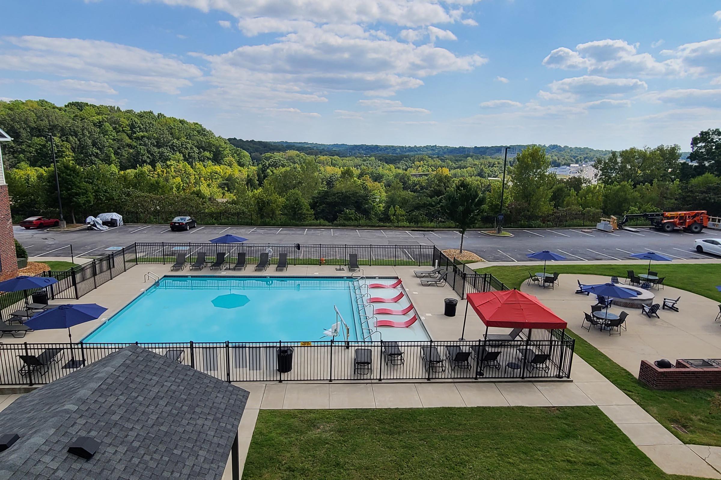 Aerial view of a pool area surrounded by green trees and hills. The pool is rectangular, with lounge chairs and umbrellas around it. There are red and white striped poolside loungers and several tables with umbrellas. The background shows a parking lot and distant hills under a partly cloudy sky.