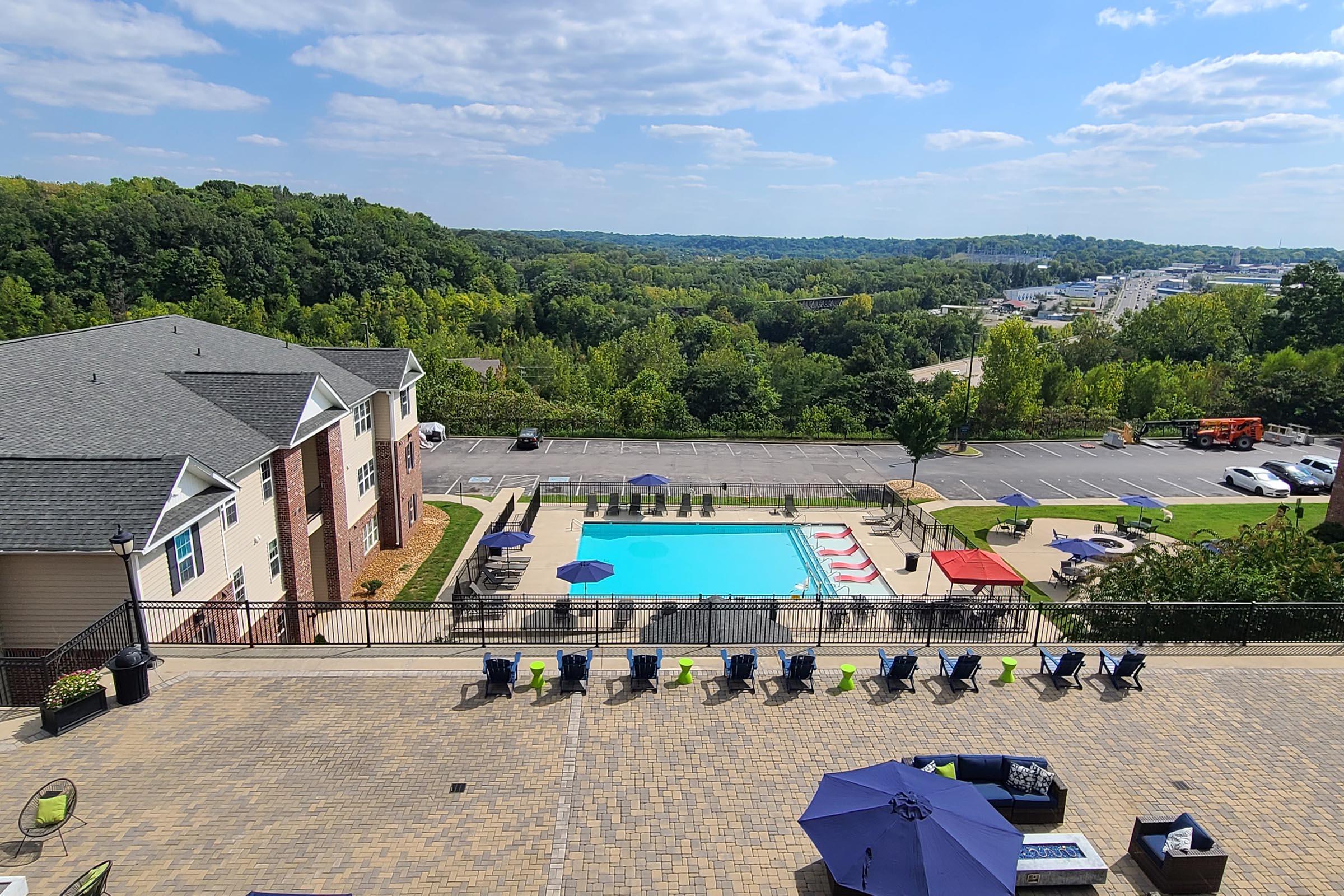 A view of a modern apartment complex overlooking a swimming pool surrounded by lounge chairs. The pool area features umbrellas and is set against a backdrop of lush green trees and a distant landscape. Bright skies and scattered clouds create a sunny atmosphere.