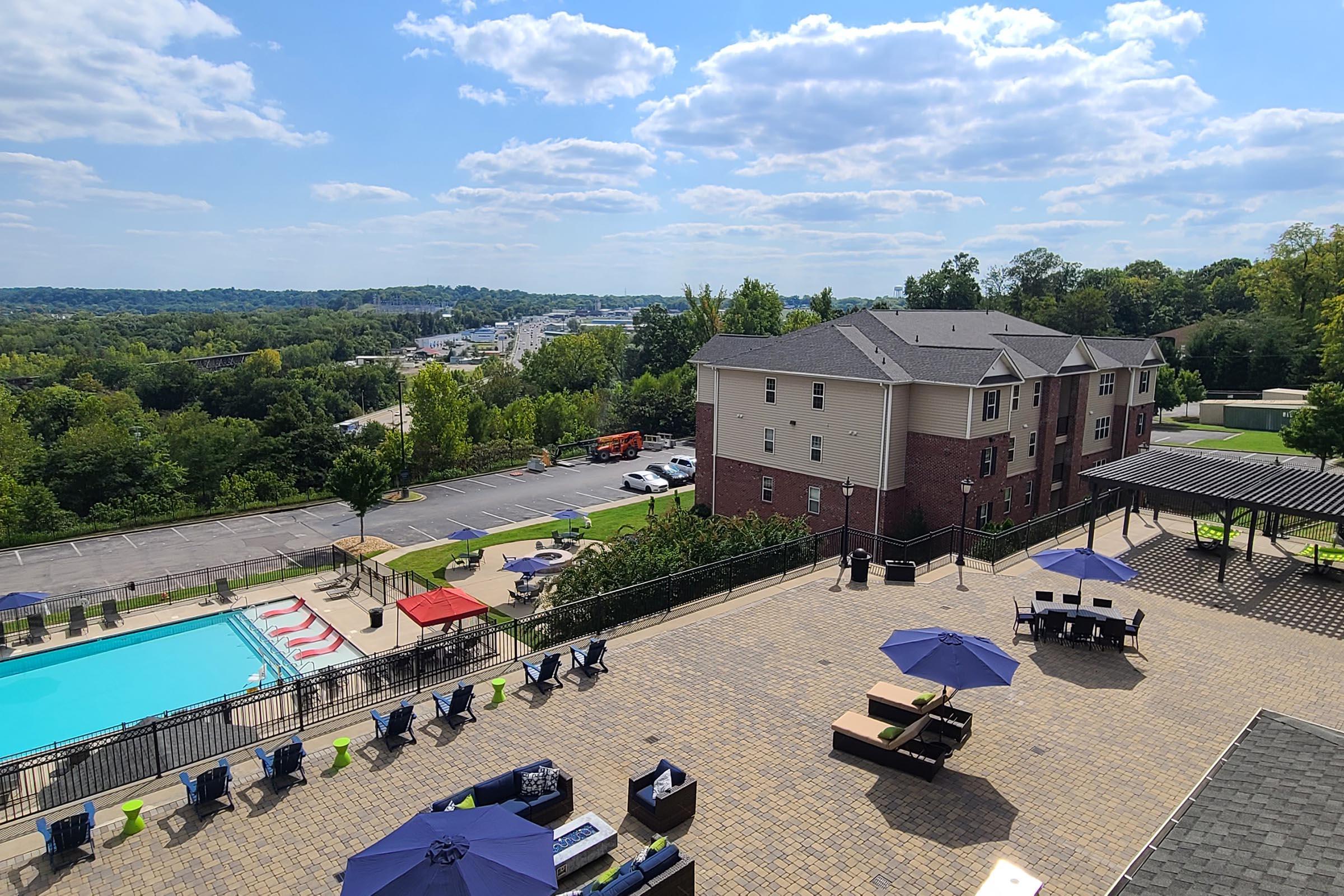 A scenic view from a high vantage point showing a swimming pool surrounded by lounge chairs and umbrellas. Nearby, there are residential buildings and a parking area, with scenic green hills and partly cloudy skies in the background. The overall atmosphere is relaxed and inviting.