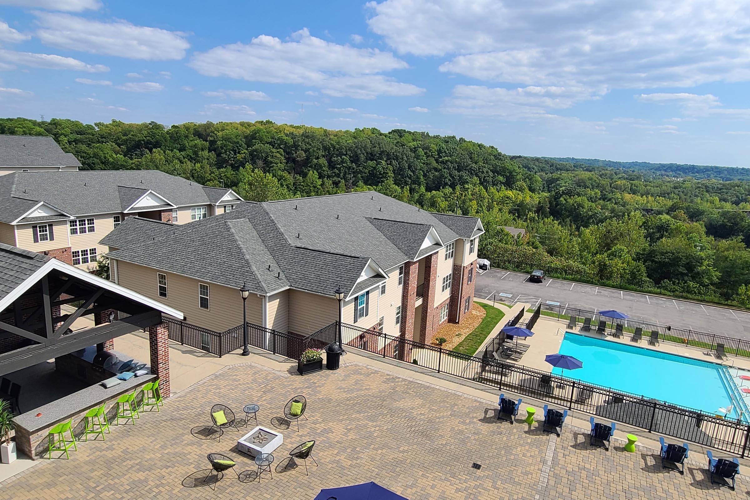 A scenic view of an apartment complex featuring multiple buildings with gray roofs, overlooking a green wooded area. A swimming pool is visible in the foreground, surrounded by lounge chairs and outdoor seating, under a partly cloudy sky.