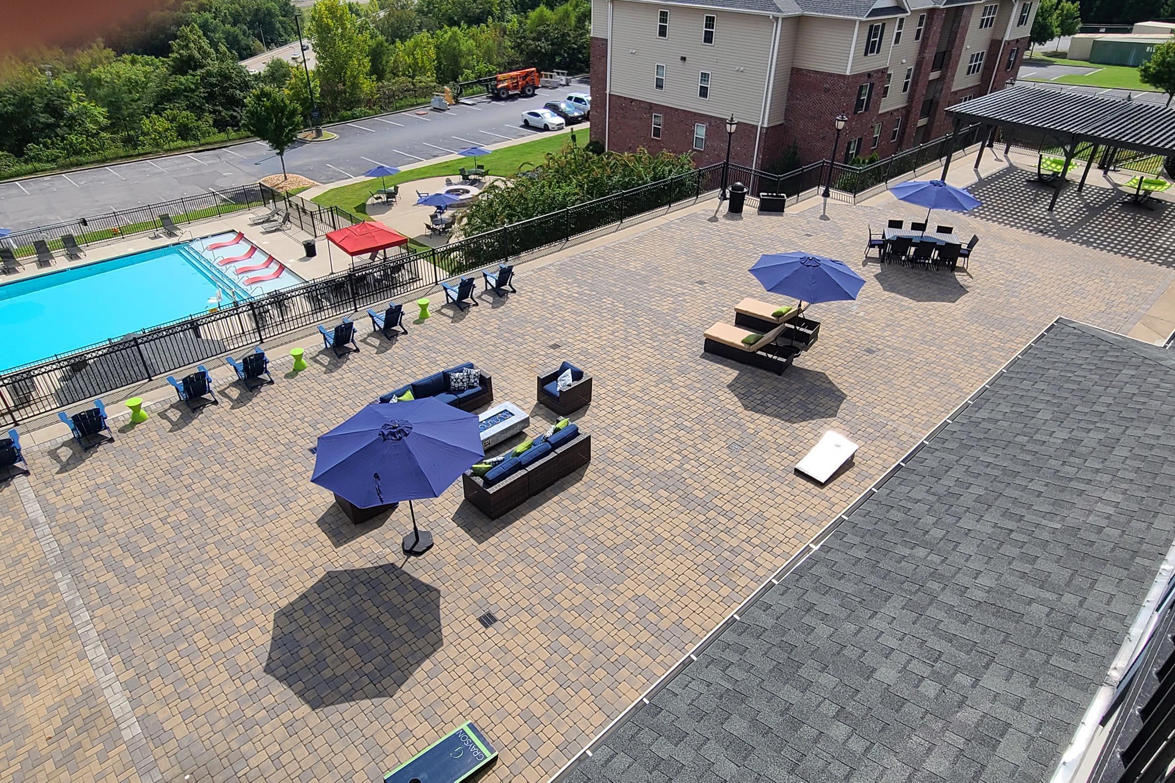 A spacious outdoor area featuring a pool, lounge chairs, and umbrellas. There are several seating arrangements with couches and tables under shaded canopies. The surrounding landscape includes greenery and residential buildings in the background, with a parking area visible.