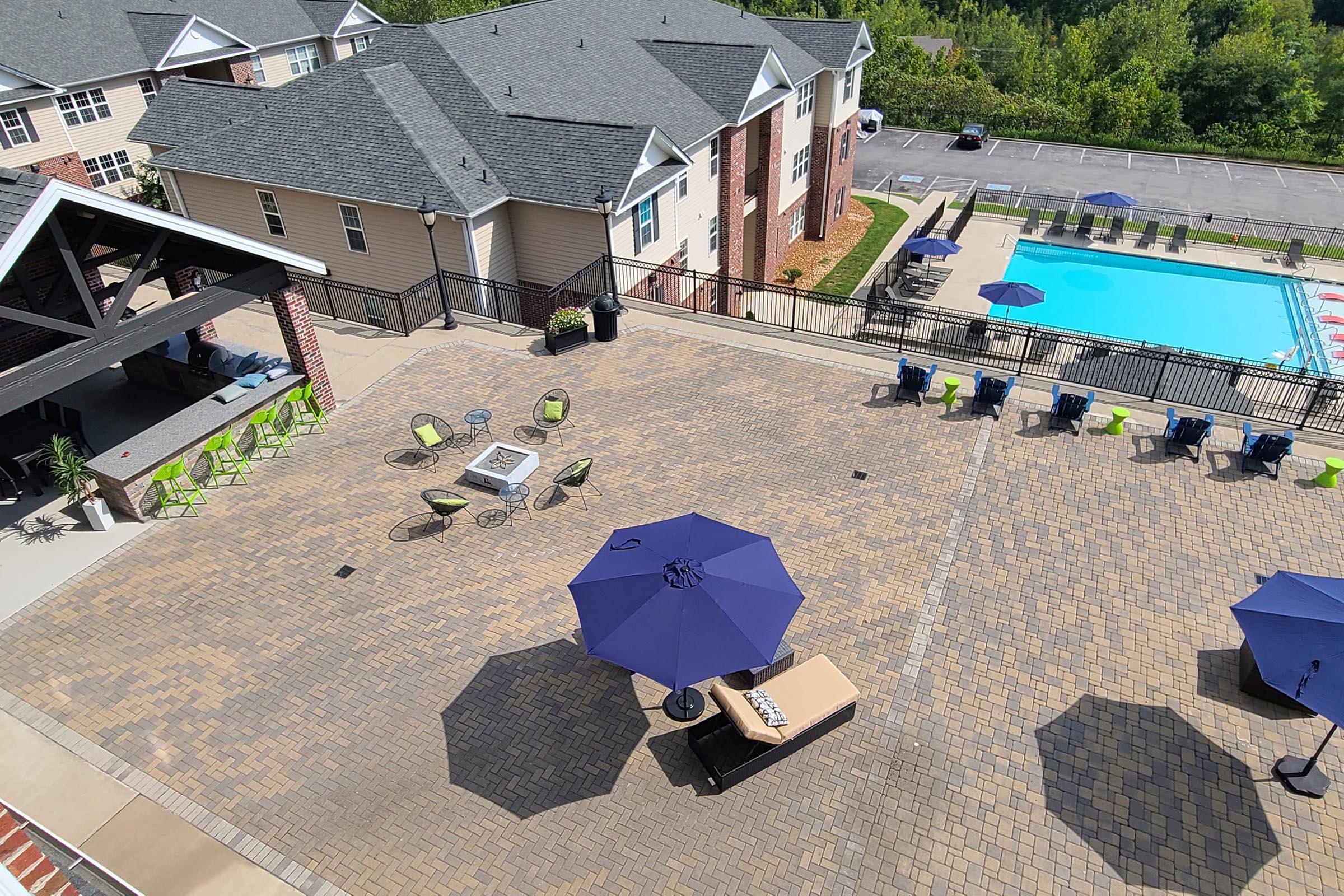 A bird's-eye view of a residential pool area featuring a large swimming pool, lounge chairs, and umbrellas. The patio is paved with square stones and includes a fire pit surrounded by seating. Nearby, there are tables and chairs on a spacious deck, with neighboring buildings visible in the background.