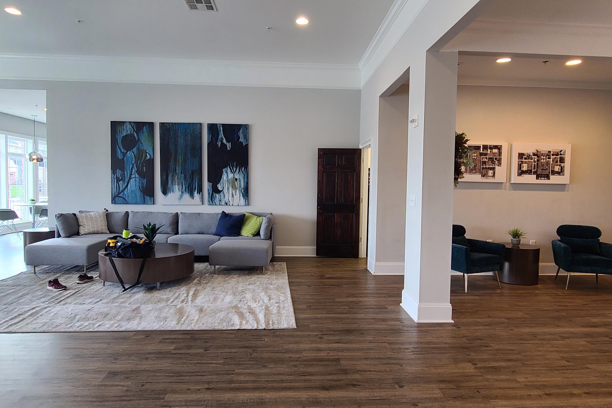 A spacious living area featuring a modern gray sectional sofa with colorful cushions, a round coffee table, and a large area rug. There are abstract paintings on the wall, a dark wooden cabinet, and two blue armchairs next to small side tables. Natural light fills the space through large windows.