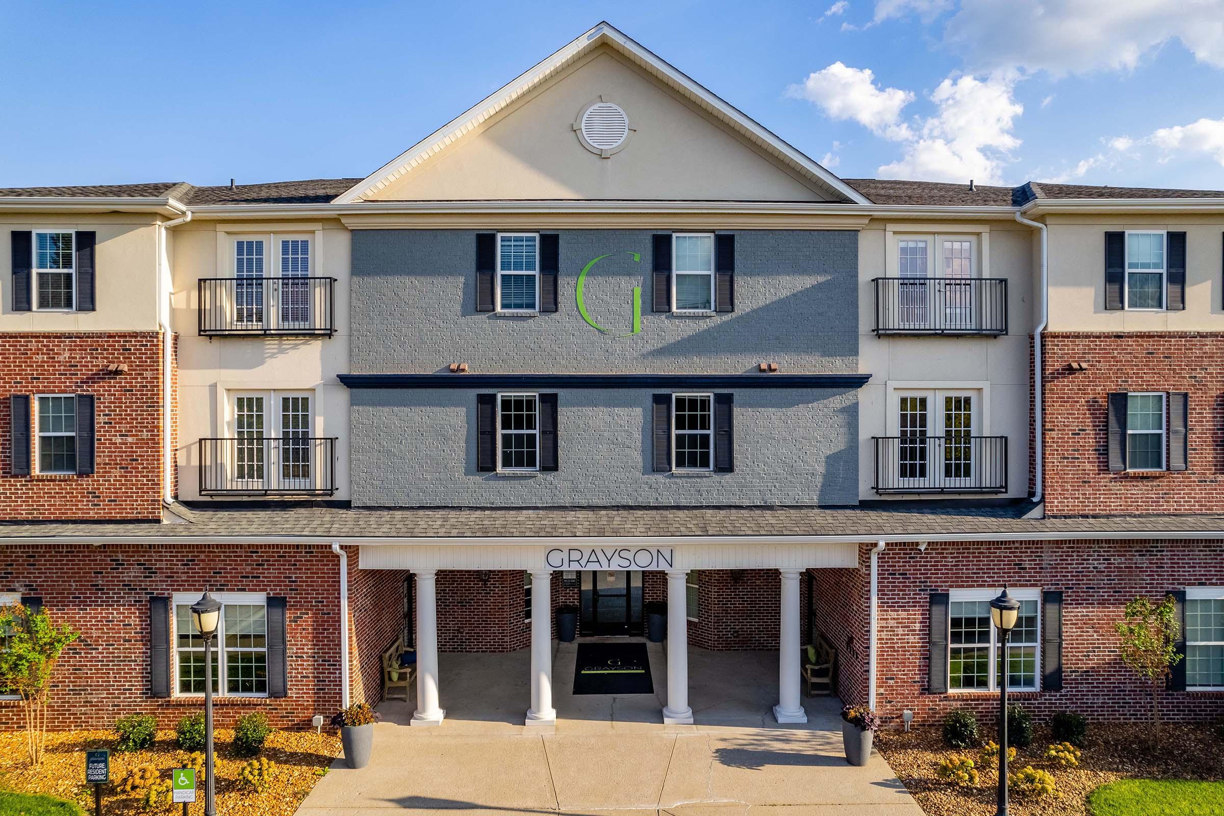 Modern brick and cream-colored building with balconies on the second floor, featuring a prominent entrance marked "GRAYSON." The facade has a mix of brick and painted surfaces, with landscaped areas and lampposts flanking the entrance. Clear blue sky and clouds in the background.