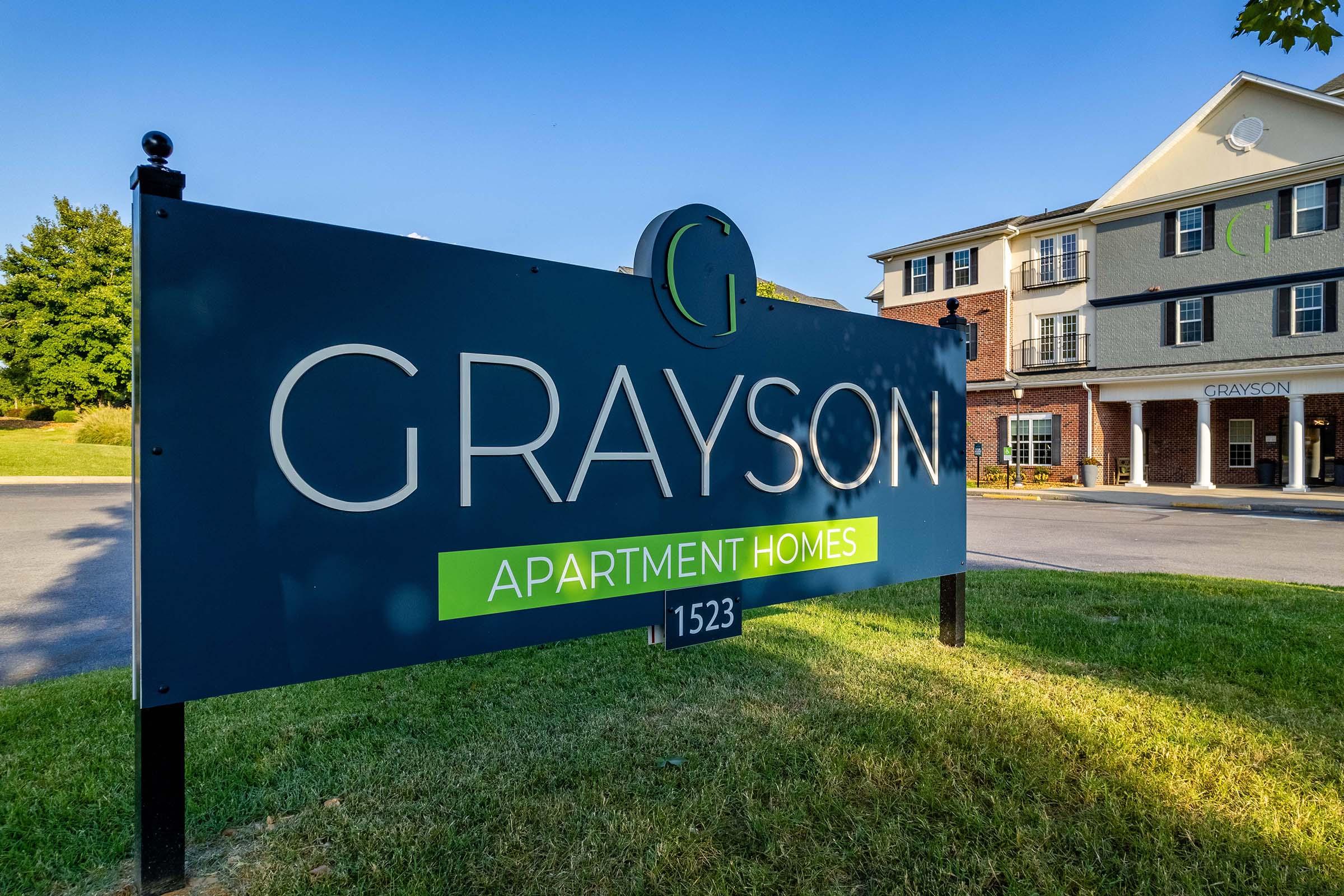 Sign for Grayson Apartment Homes, featuring the text "GRAYSON" prominently displayed in large letters, along with "APARTMENT HOMES" in a smaller font. The sign is set against a clear blue sky with greenery in the background. A building can be seen nearby, indicating it's part of the apartment complex.