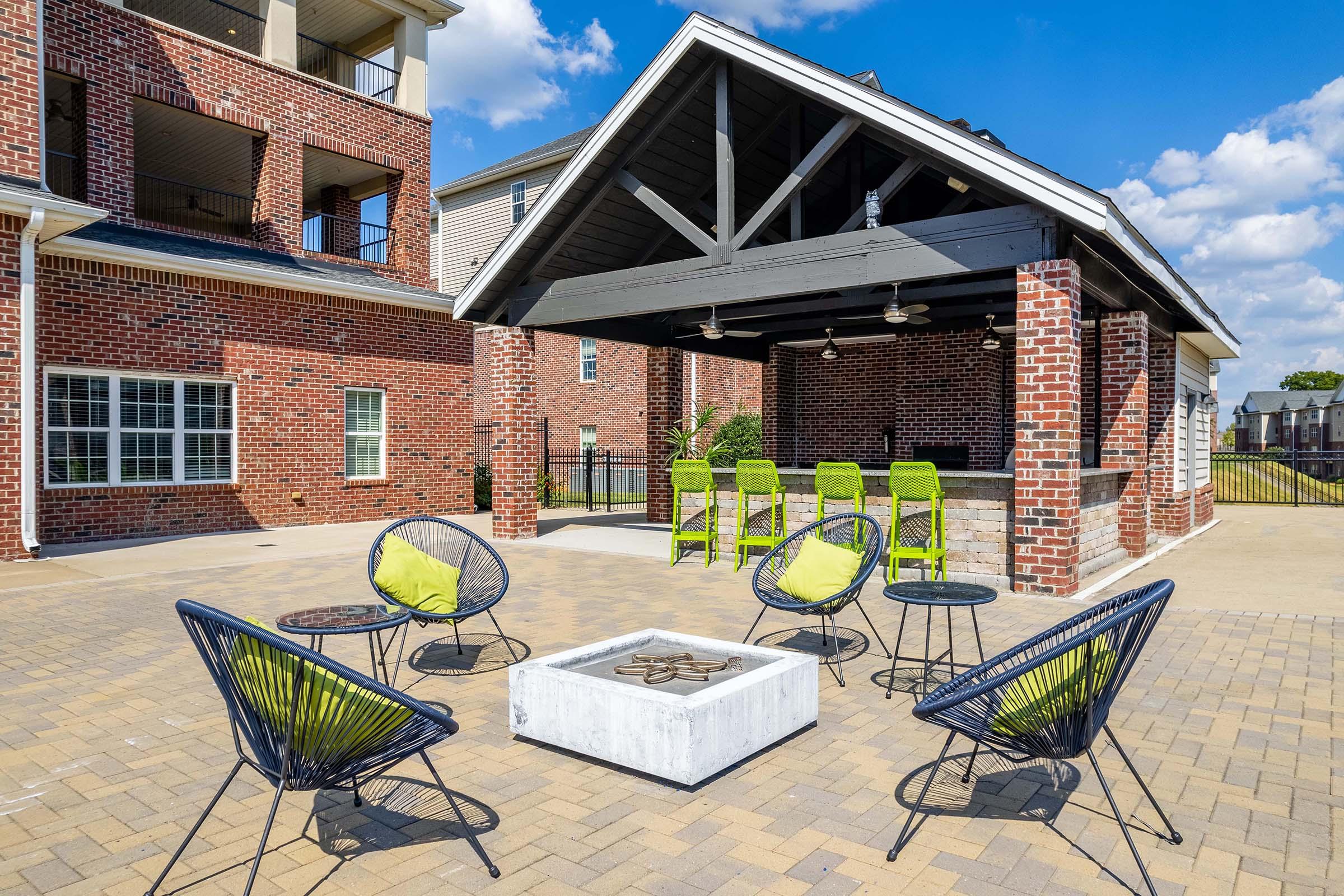 Outdoor lounge area featuring a covered pavilion with a bar, surrounded by six black and green chairs arranged around a modern fire pit. The setting includes brick architecture and a clear blue sky with scattered clouds.
