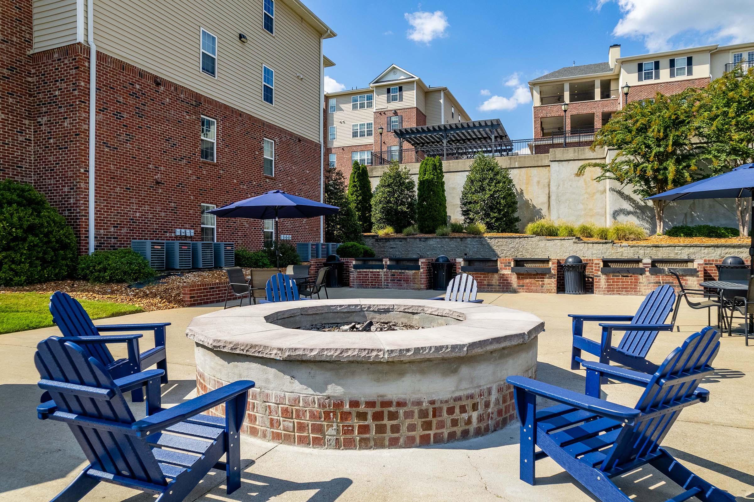 A communal fire pit surrounded by blue adirondack chairs in a landscaped outdoor area. In the background, modern apartment buildings and shaded umbrellas are visible, creating a relaxed atmosphere. The scene is set on a sunny day with a clear blue sky.
