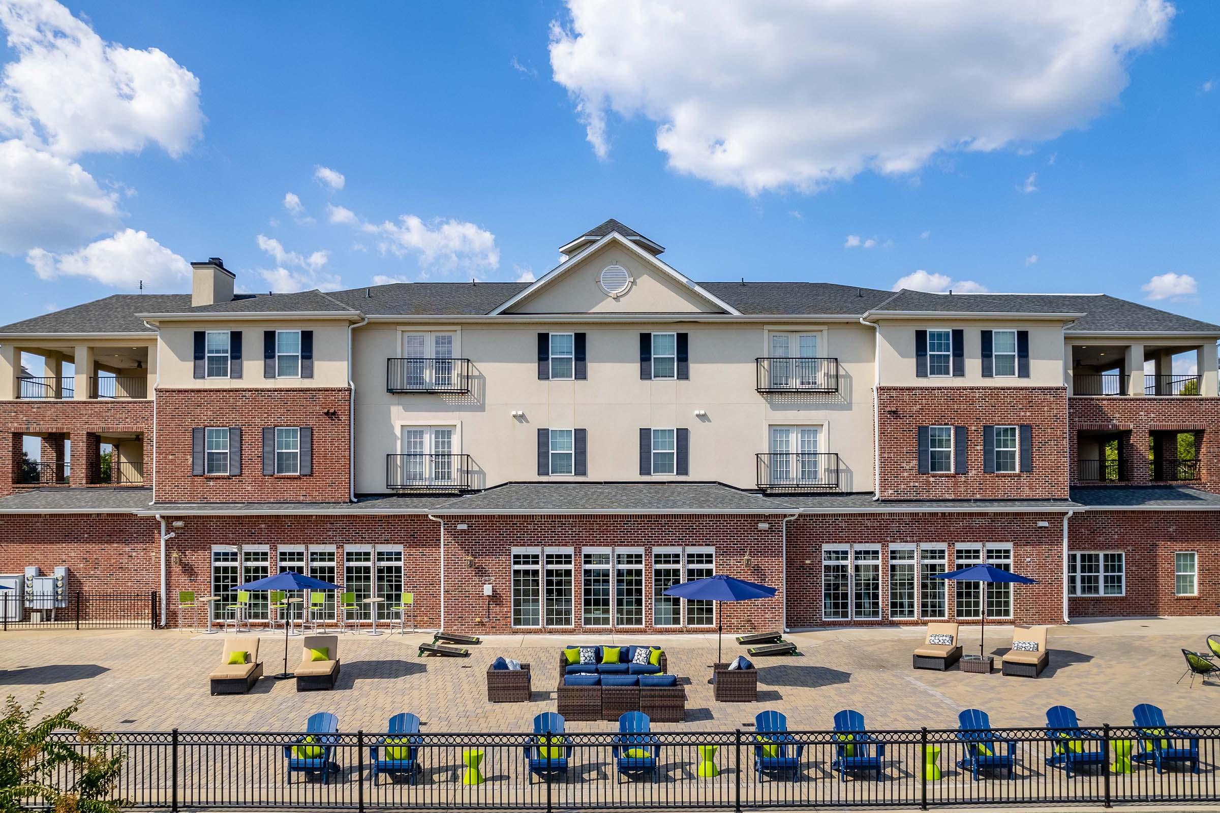 A modern building with a multi-story structure featuring a combination of brick and light-colored exterior. The foreground includes a patio area with lounge chairs, tables, and umbrellas. Clear blue sky and fluffy clouds are visible in the background, suggesting a bright, sunny day.