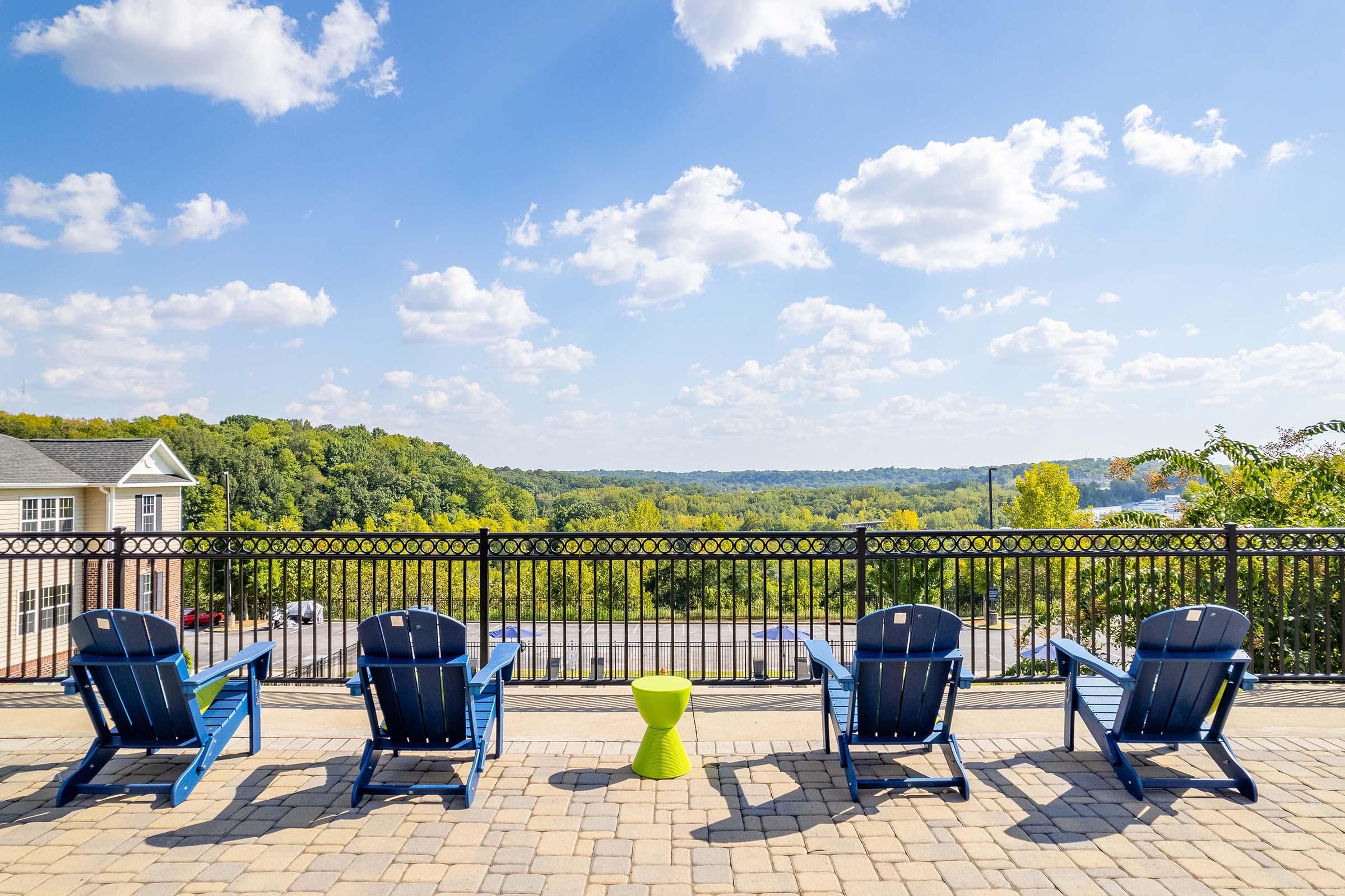 A scenic outdoor view featuring four blue Adirondack chairs arranged on a patio. In the background, lush green hills stretch under a bright blue sky dotted with fluffy white clouds. A small green table sits between the chairs, offering a cozy spot to enjoy the landscape.