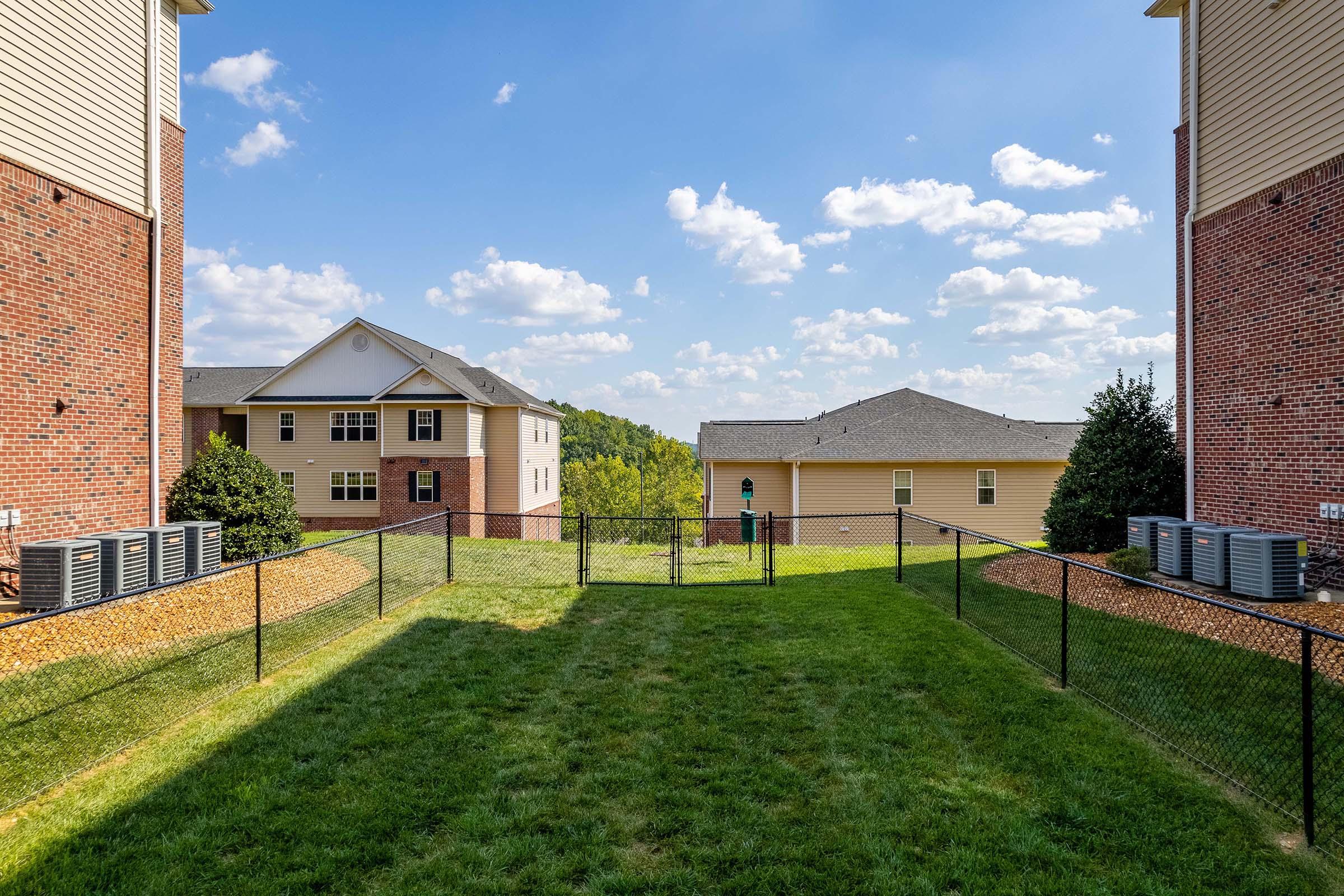View of a fenced grassy area between two residential buildings. One building features a brick facade, while the other has a light-colored exterior. In the background, there are rolling hills and a partly cloudy blue sky. Air conditioning units are visible along the side of the buildings.
