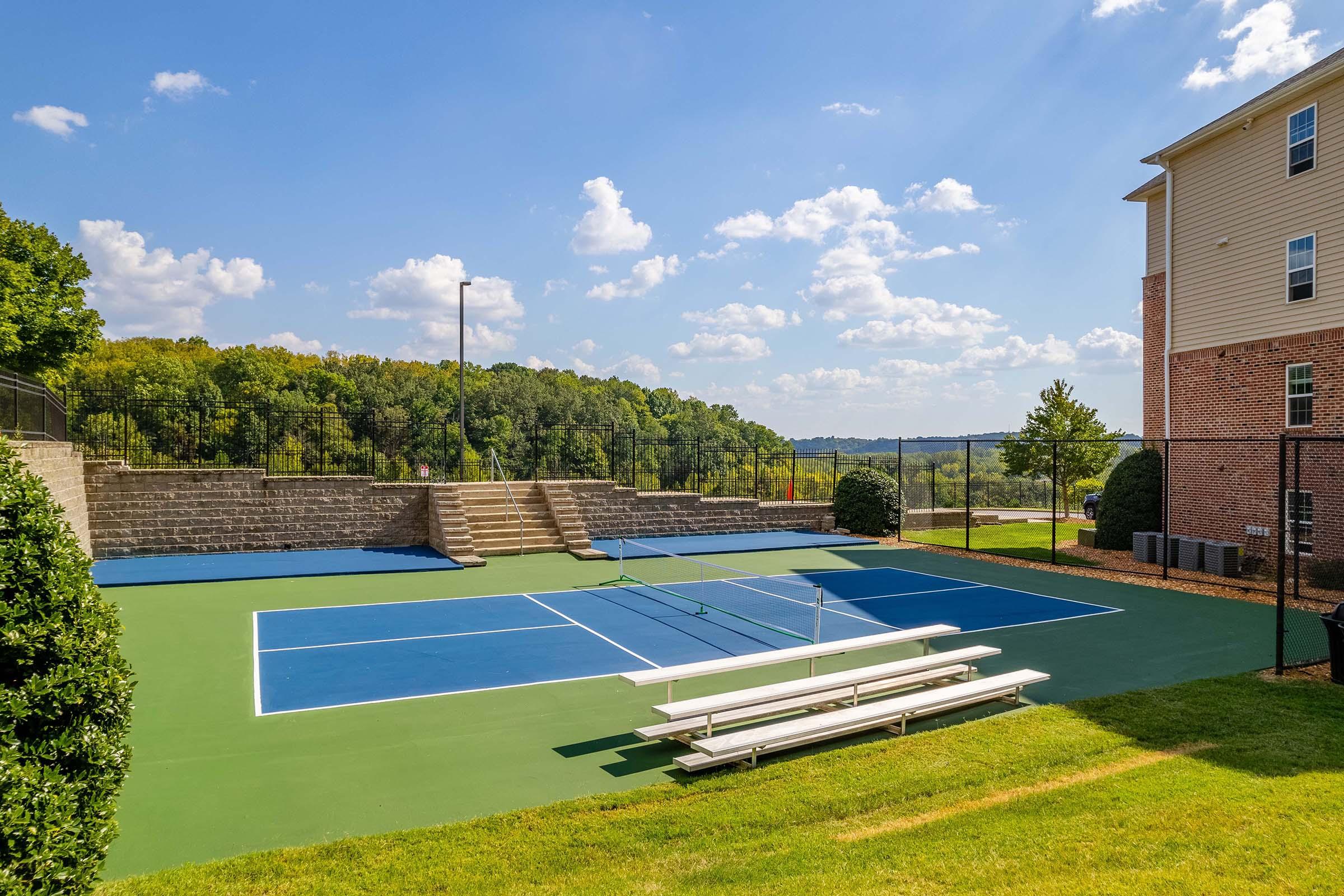 A sunny outdoor area featuring two tennis courts with blue surfaces and white lines. There are benches for spectators nearby, surrounded by greenery and trees in the background. A residential building is visible on the right side. The sky is partly cloudy, adding to the scenic view.