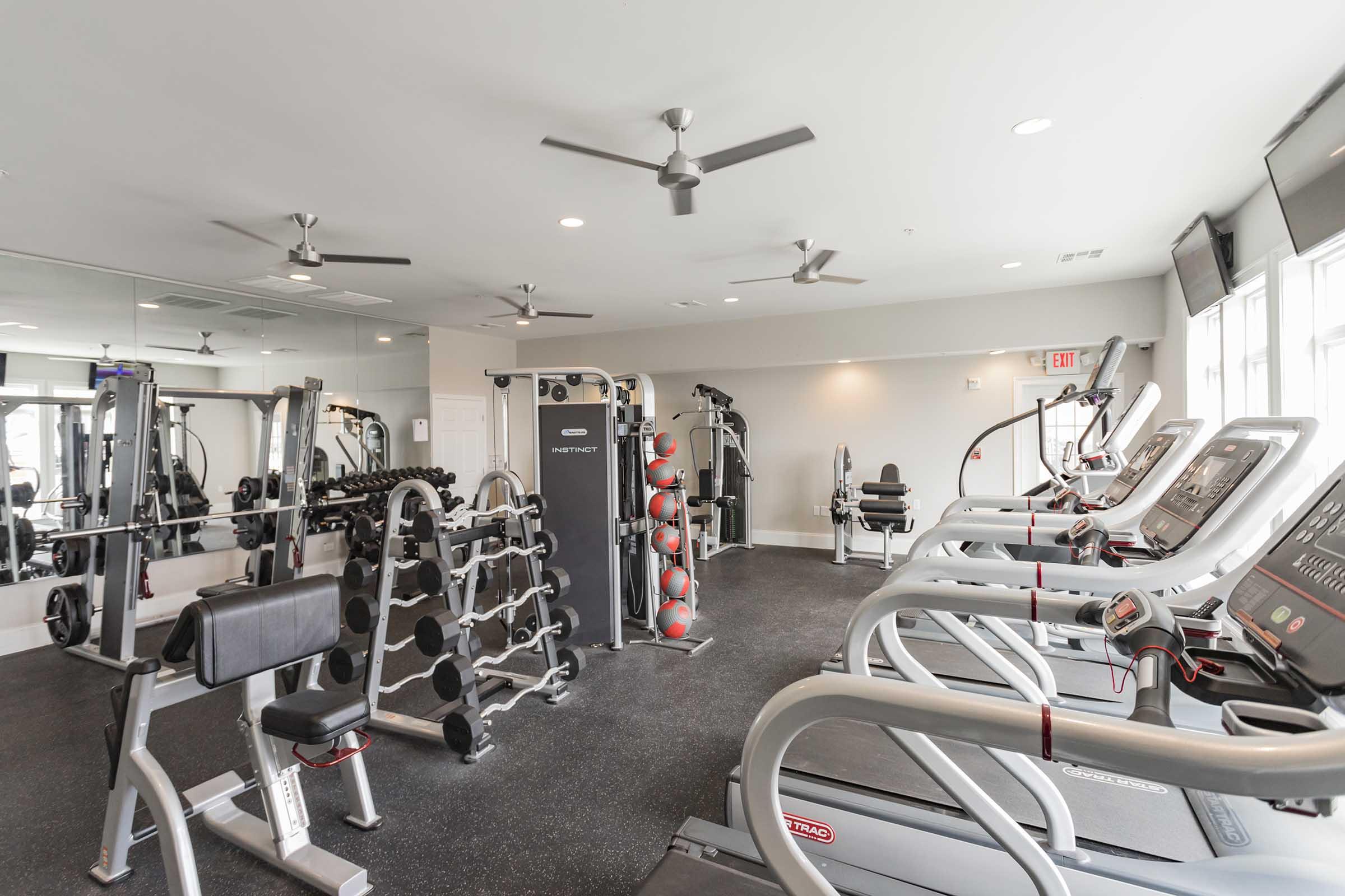 A well-equipped gym featuring various exercise machines including treadmills, weight benches, and free weights. The space is brightly lit with ceiling fans and mirrors, creating an open and inviting atmosphere for workouts.