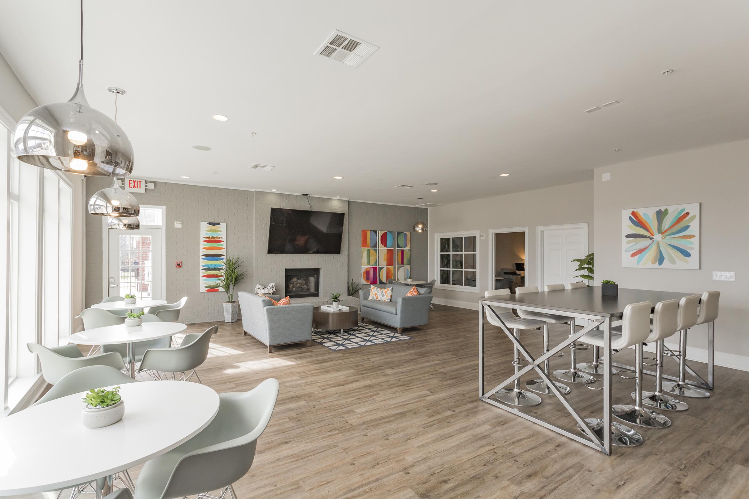 Modern lounge area featuring a mix of seating options, including chairs and a large table. Large windows allow natural light in, and colorful artwork adorns the walls. A cozy fireplace is centered in the space, with a stylish, contemporary design and warm wood flooring throughout.