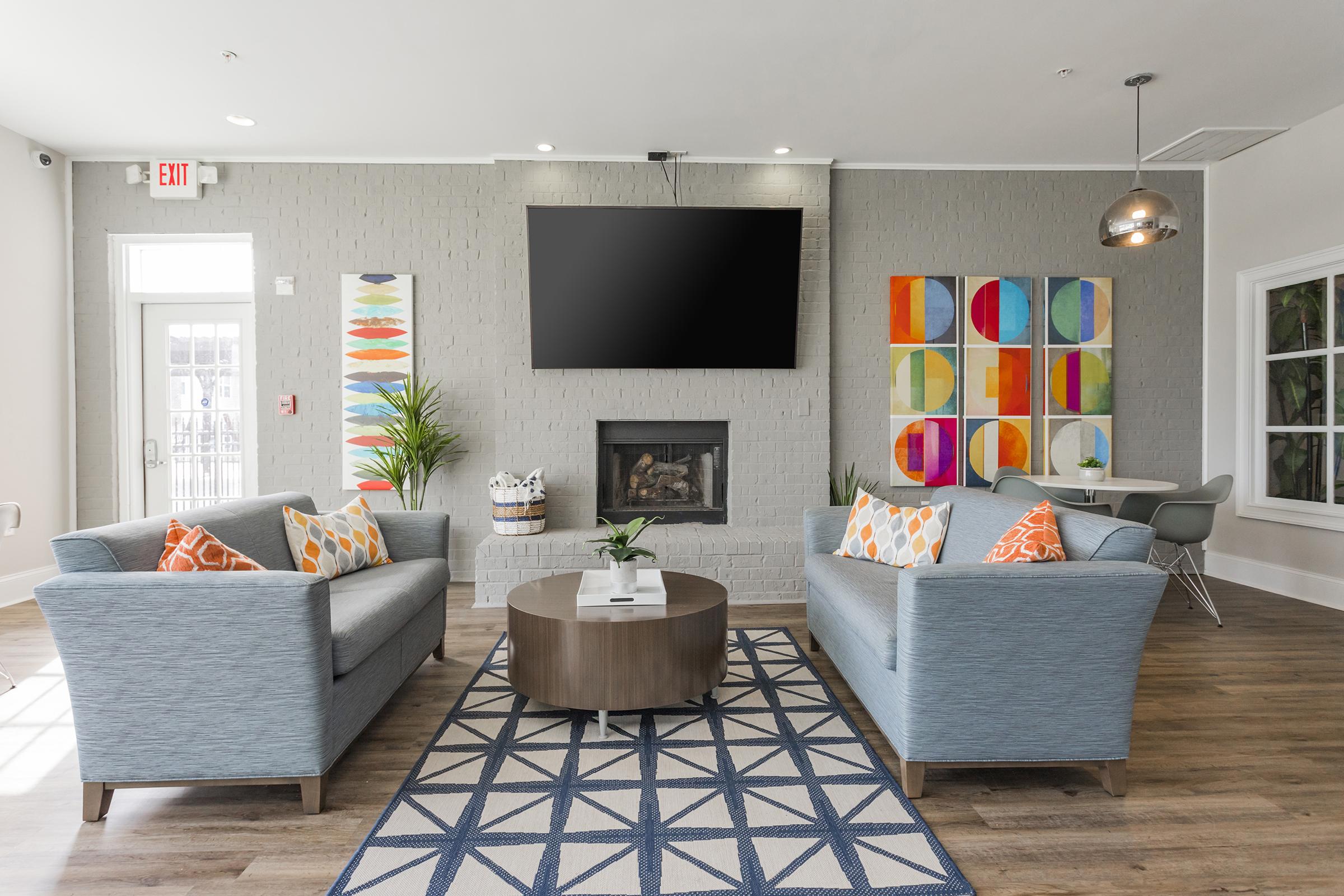 A modern living room with two light blue sofas adorned with colorful pillows. A circular coffee table sits in the center on a geometric patterned rug. The walls feature a flat-screen TV, a fireplace, and vibrant artwork. Large windows allow natural light, and potted plants add a touch of greenery.
