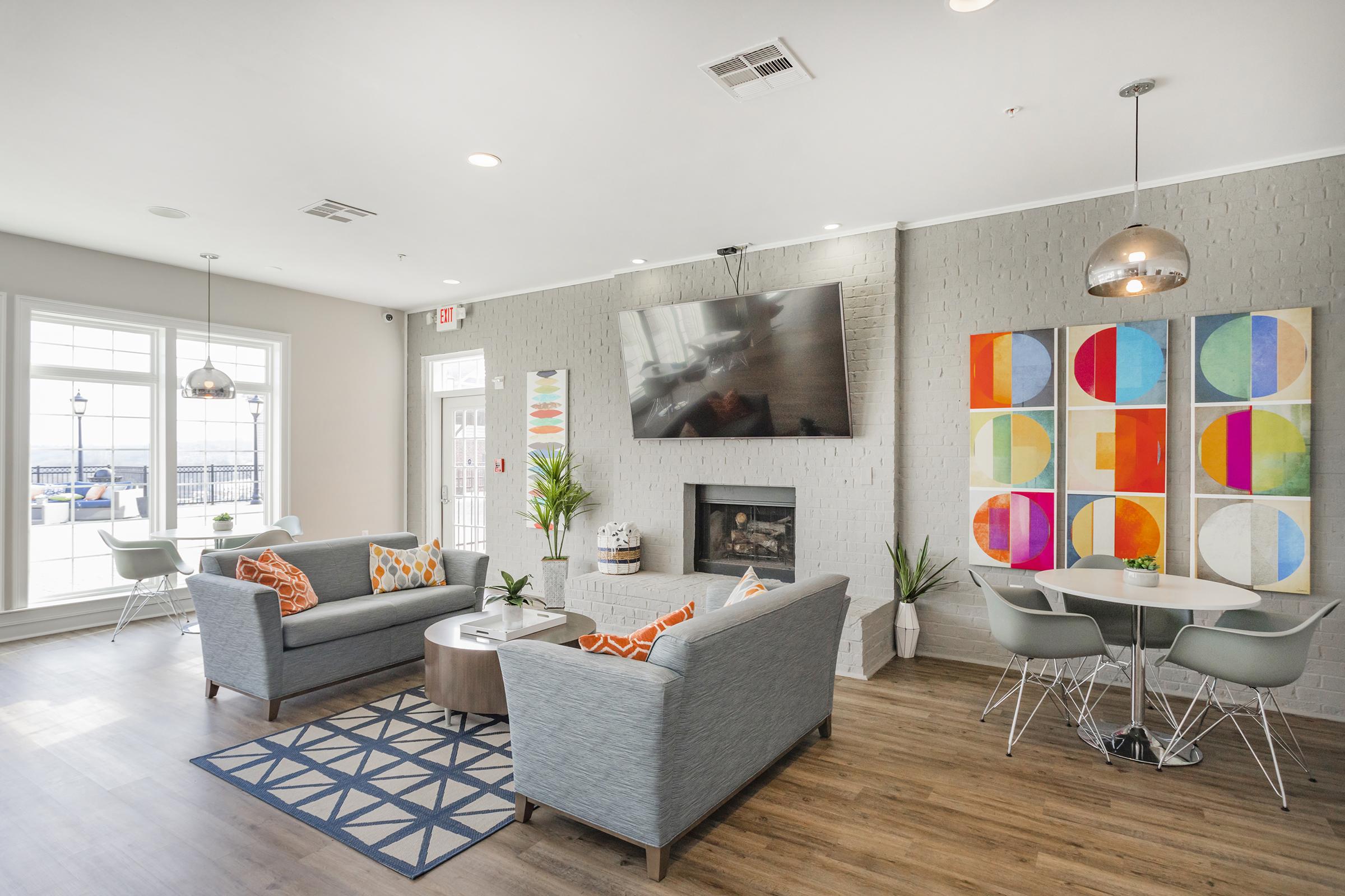 A modern living area featuring two light blue sofas with orange and white cushions, a fireplace, a large TV mounted on the wall, and colorful abstract wall art. A geometric-patterned rug complements the space, while round dining tables and chairs add to the contemporary design. Large windows let in natural light.