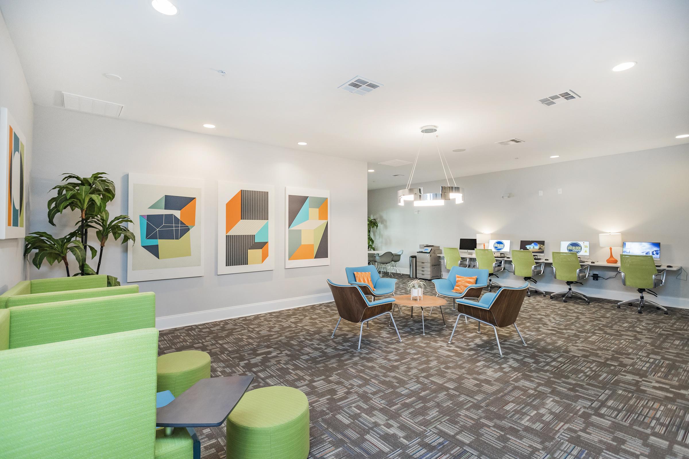 A modern office space featuring two types of seating: green, upholstered chairs and wooden lounge chairs. The walls are adorned with colorful geometric artwork. There is a central coffee table, and behind it, two workstations with computers are visible. Bright lighting enhances the contemporary atmosphere.