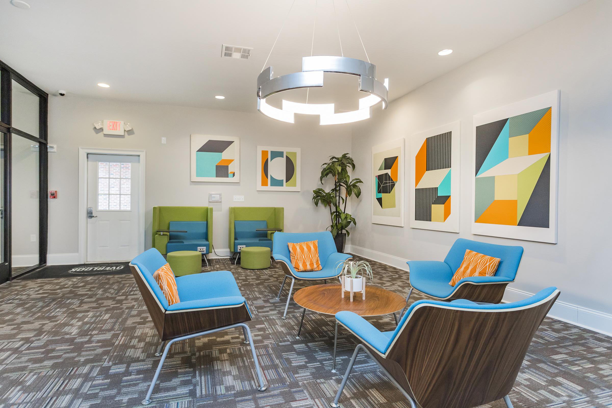 A modern lobby area featuring contemporary seating in bright colors of blue and orange, a round wooden table, and abstract artwork on the walls. There are green lounge chairs and decorative plants, creating a welcoming and stylish atmosphere. The space has neutral walls and a patterned floor.