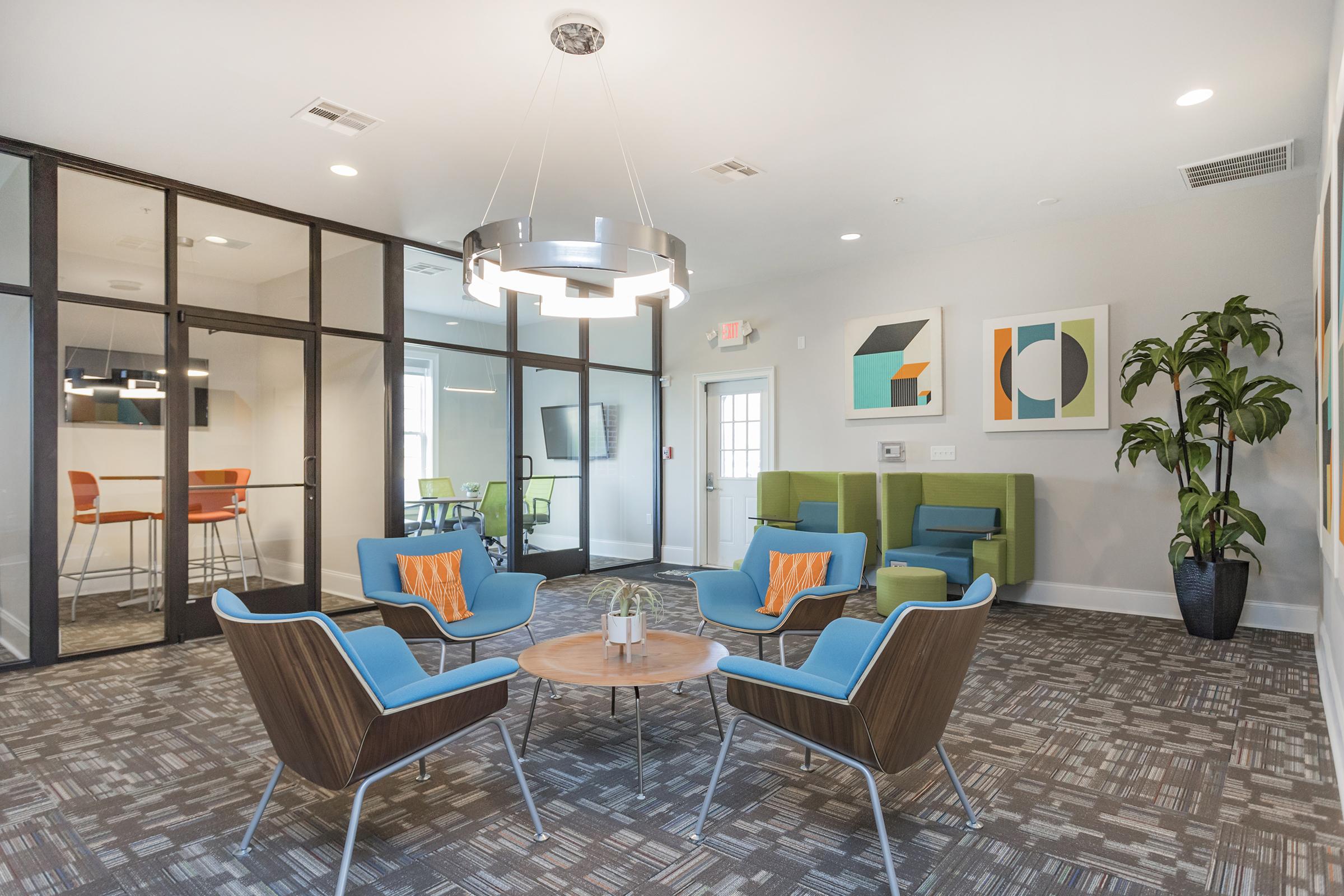 A modern lounge area featuring blue and green chairs, a round wooden table, and a stylish light fixture. There are framed abstract art pieces on the walls, a potted plant in the corner, and a glass-walled meeting room in the background. The decor creates a welcoming and contemporary atmosphere.