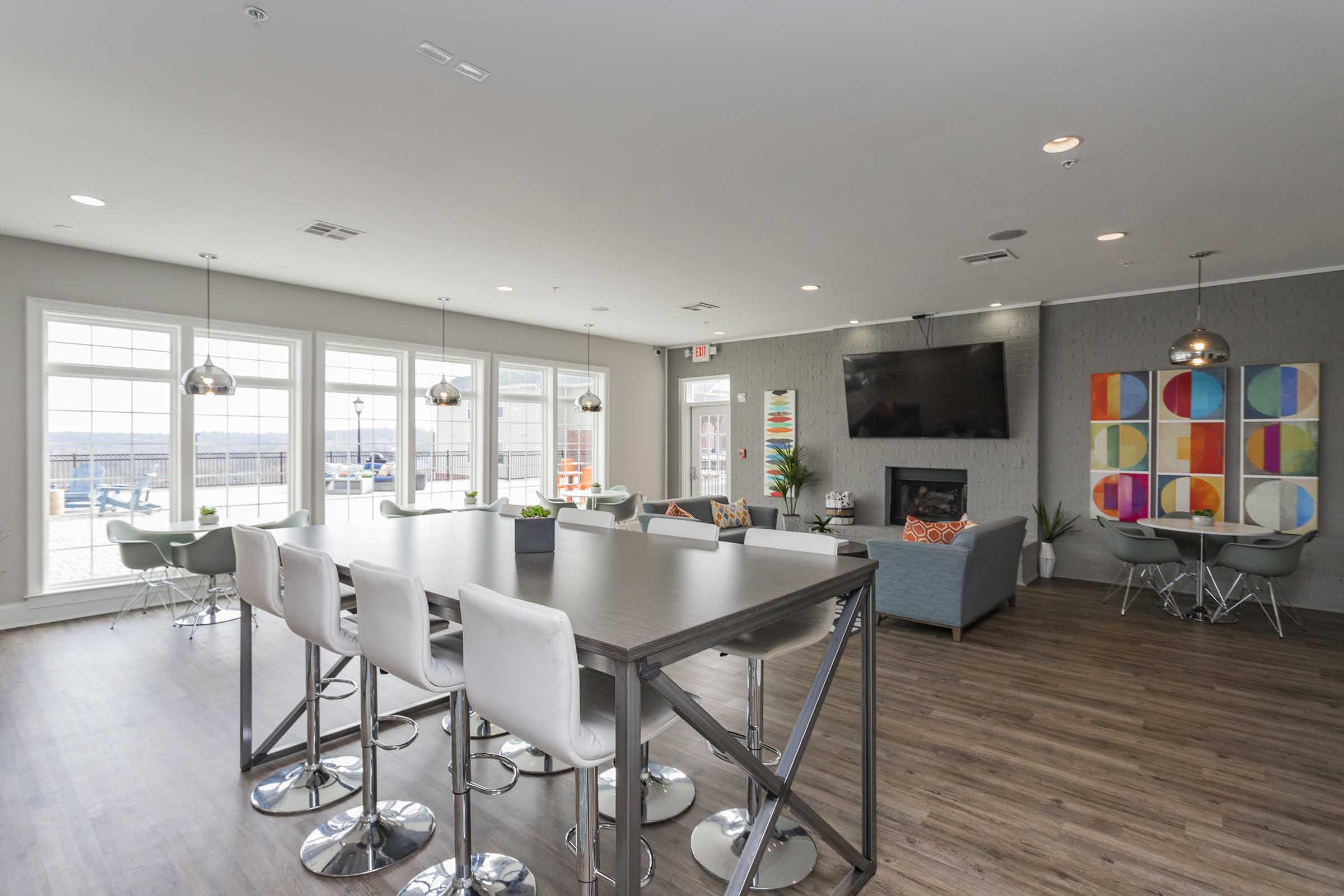 A modern lounge area featuring a gray sofa, a large flat-screen TV on the wall, and a high table with white bar stools. Large windows allow natural light and offer a view of the outside. Colorful wall art and plants add a vibrant touch to the space. The flooring is a warm wood finish.