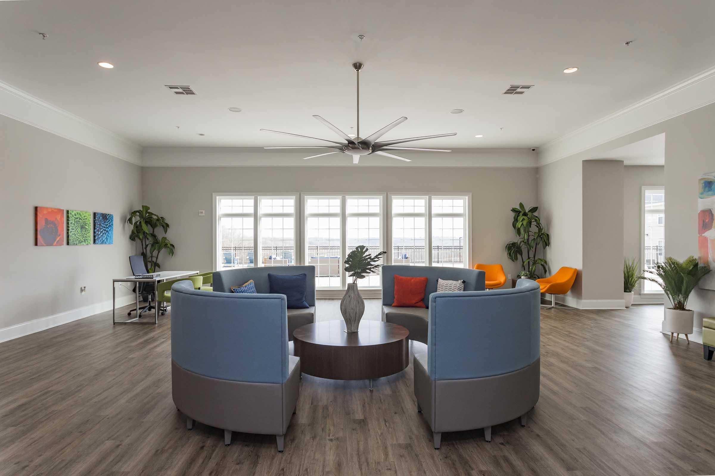 A spacious lounge area featuring two modern blue armchairs and a round wooden coffee table at the center. Colorful wall art adorns the light gray walls, with large windows providing natural light. Comfortable seating includes orange and green chairs, along with potted plants enhancing the inviting atmosphere.