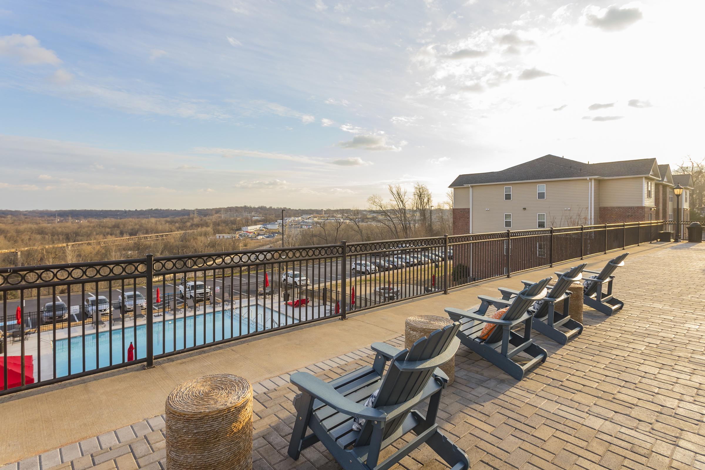 A tranquil outdoor scene featuring a lined-up row of blue adirondack chairs overlooking a swimming pool and scenic landscape. The area is bathed in warm sunlight, with a walled balcony providing a view of trees and buildings in the distance.