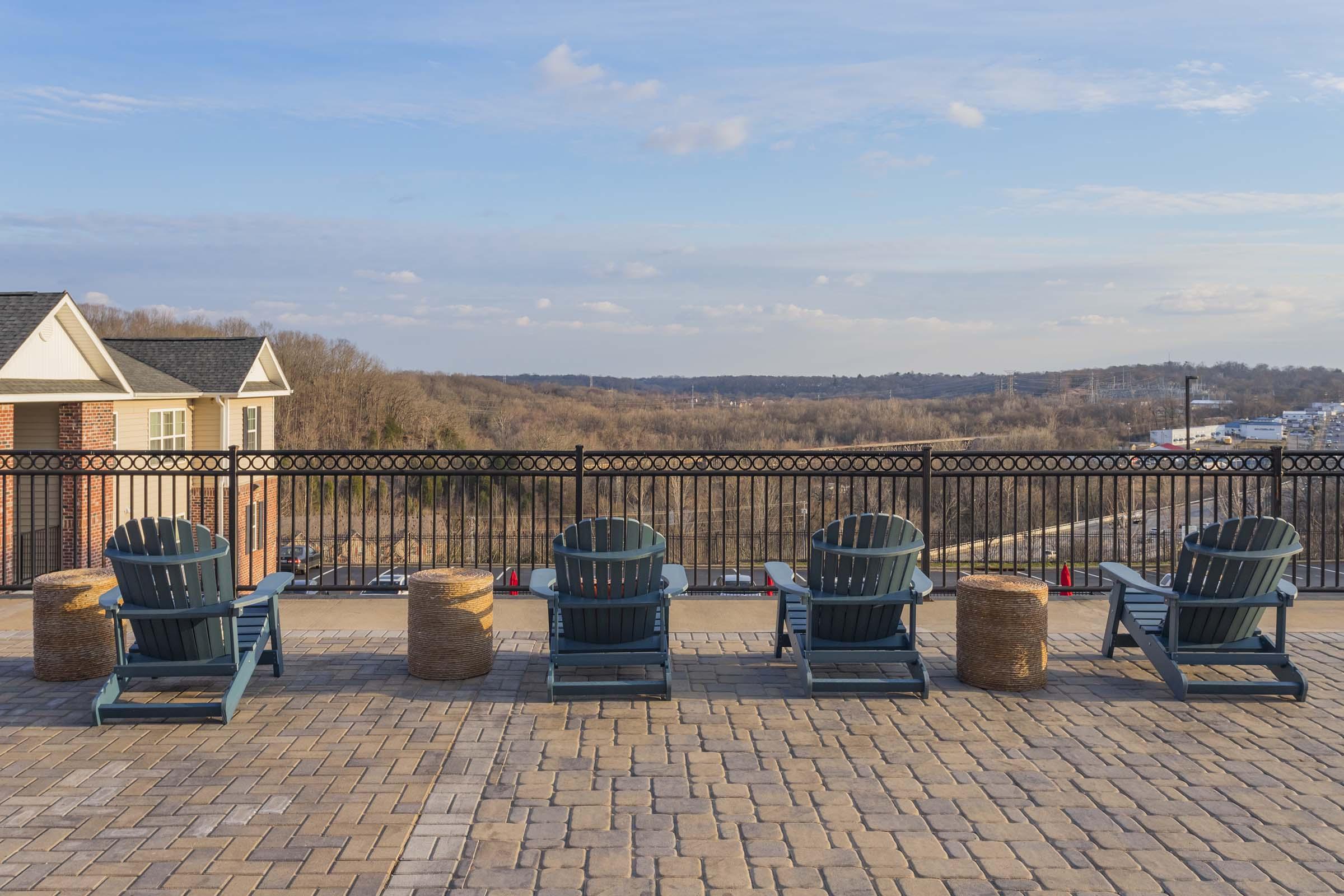 Four blue Adirondack chairs are lined up along a balcony, overlooking a scenic view of rolling hills and a valley. The area is paved with stone and features decorative round straw bales. A black iron fence adds a stylish touch, while the sky is clear with a few clouds.