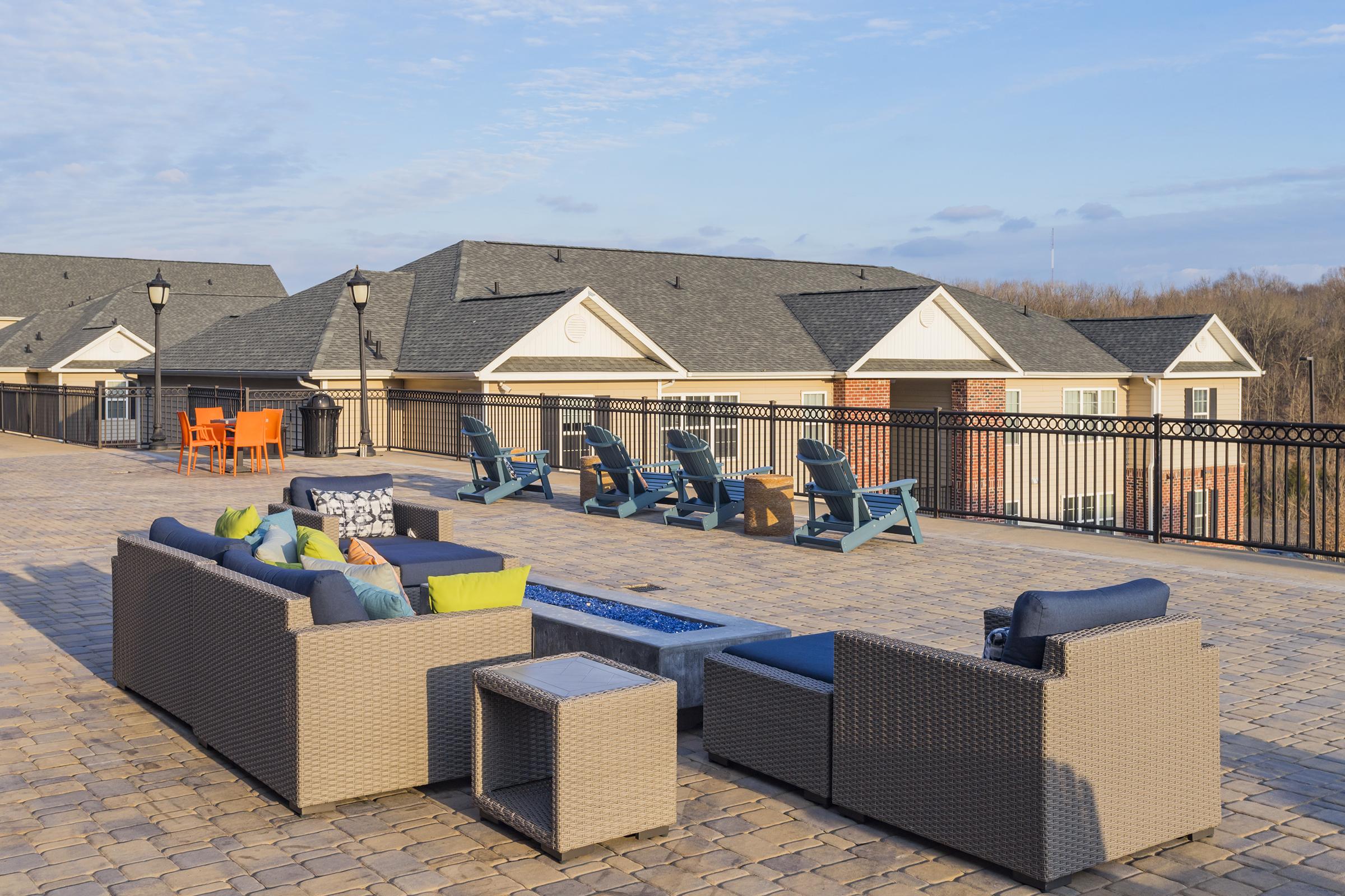A rooftop terrace featuring stylish outdoor furniture, including a sectional sofa with colorful cushions, a coffee table, and several lounge chairs. The area is surrounded by a fence and offers a scenic view of nearby buildings and the sky, creating a relaxing atmosphere for gatherings.