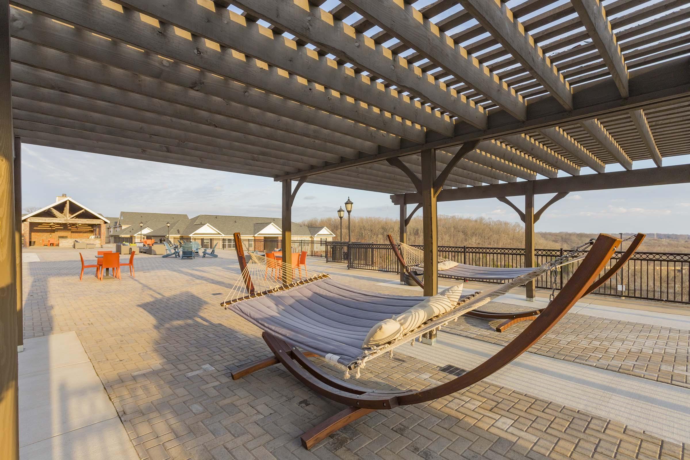 A spacious outdoor patio featuring a wooden pergola, with a hammock and several orange chairs scattered around. The area overlooks a scenic landscape, and there are additional buildings in the background, creating a relaxing retreat atmosphere. Soft sunlight adds warmth to the scene.