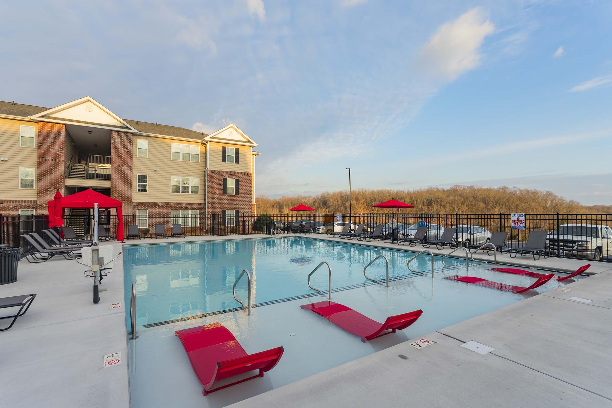 A clear pool area featuring a large swimming pool with red lounge chairs and umbrellas. Surrounding the pool are several deck chairs and a fenced perimeter, with a multi-story apartment building in the background against a blue sky.