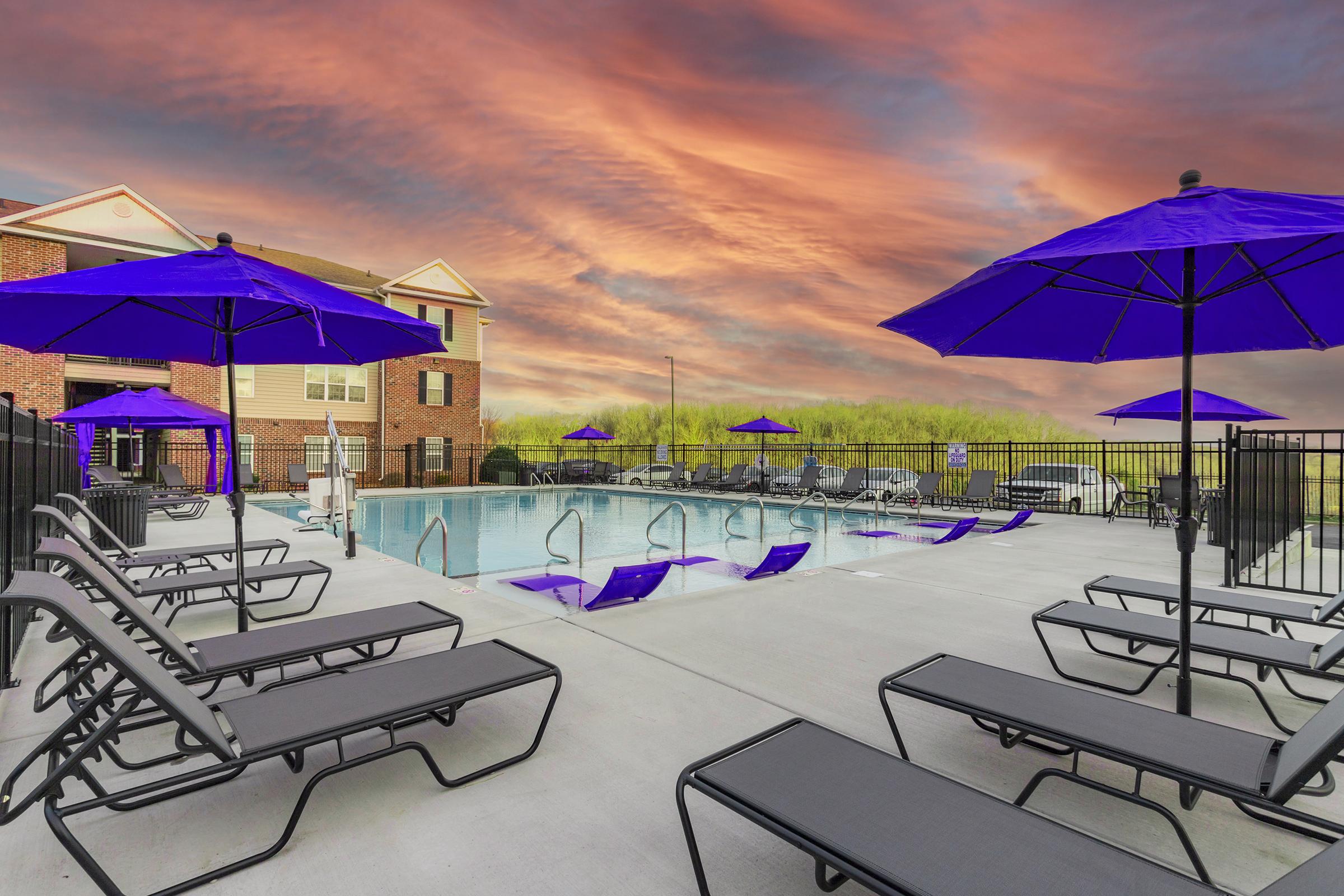 A vibrant outdoor swimming pool area featuring purple umbrellas and lounge chairs. The pool is surrounded by a black fence, with a backdrop of a colorful sunset sky. Nearby, there are additional chairs and a well-maintained landscape, creating a relaxing ambiance.