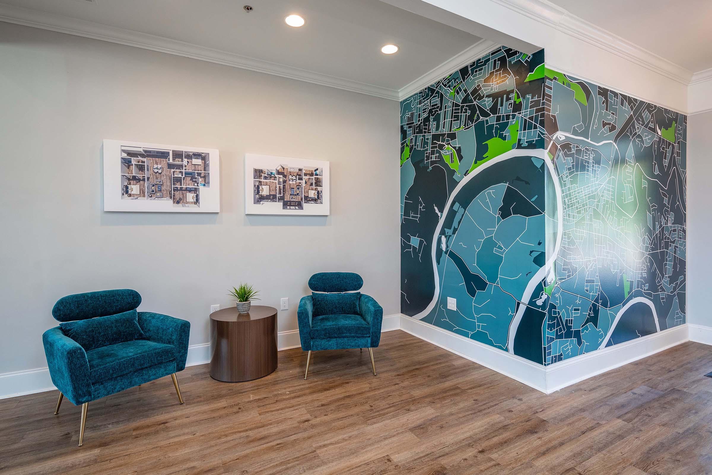 A modern interior space featuring two teal armchairs beside a small circular table. On the wall, there are two framed artworks and a large mural displaying a stylized map with shades of blue and green. The wooden floor adds warmth to the sleek design. Natural light illuminates the area.