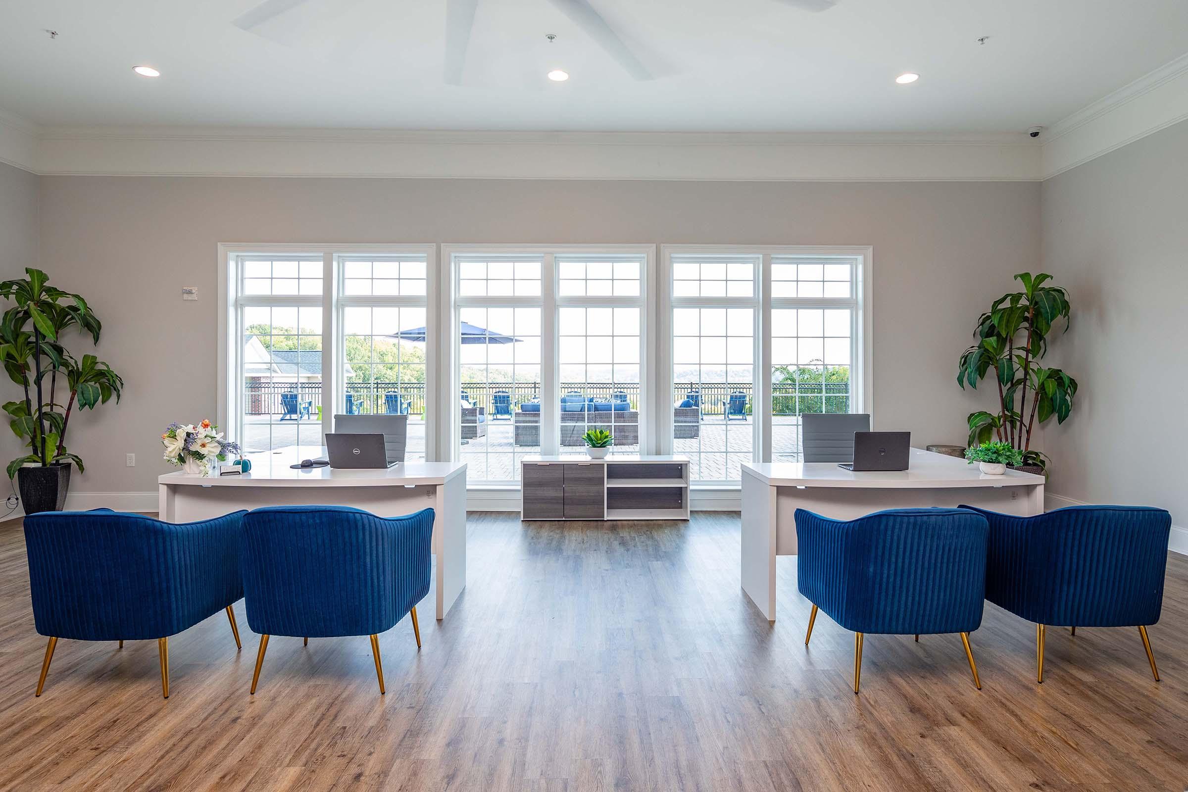A modern office space featuring two desks with laptops, surrounded by blue upholstered chairs. Large windows provide a view of an outdoor patio, and potted plants add greenery to the room. The floor is wooden, creating a warm, inviting atmosphere.
