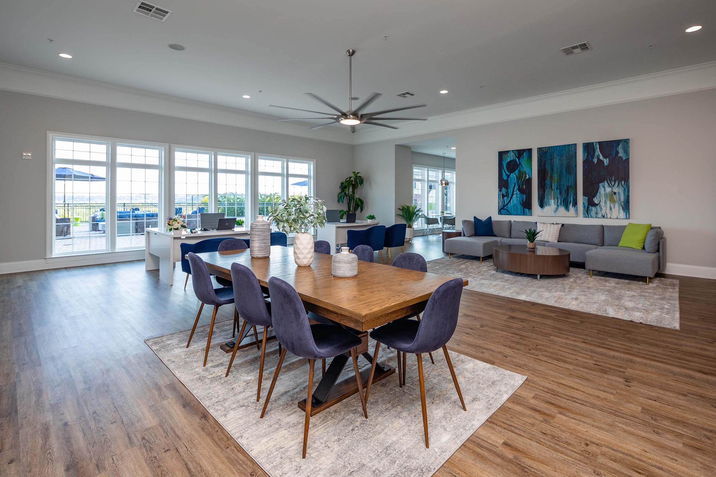 A spacious modern lounge area with large windows offering natural light. It features a wooden dining table surrounded by navy blue chairs, a gray sofa, and a decorative rug. Two abstract paintings adorn the wall, complemented by potted plants, creating a welcoming atmosphere.