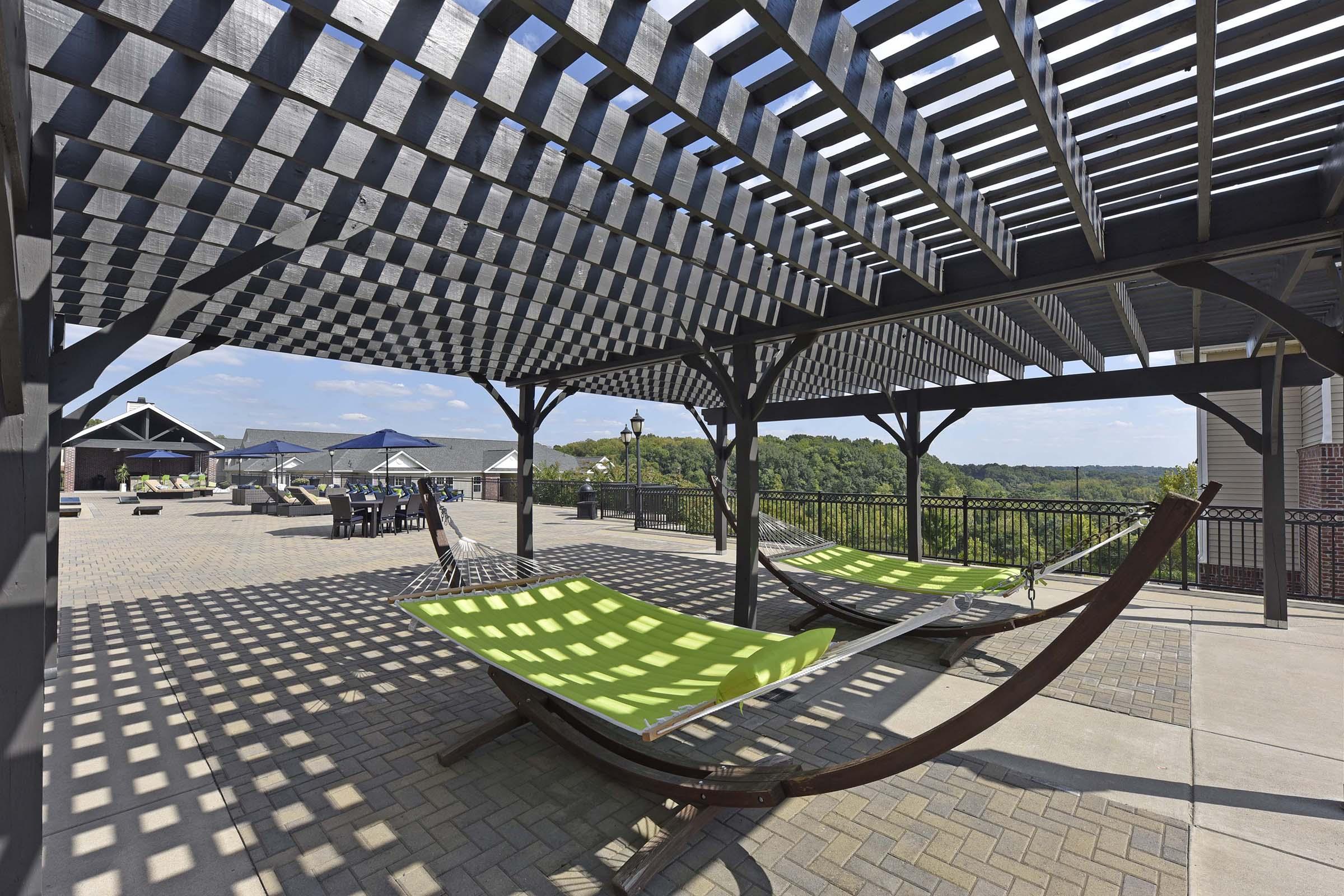 A spacious outdoor patio area featuring two green hammocks under a wooden pergola with a striped pattern. The background shows a scenic view of trees and blue skies, with seating areas and umbrellas in the distance, creating a relaxing atmosphere.