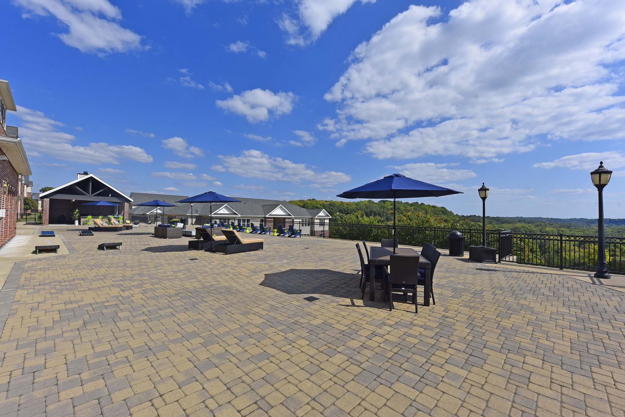 An outdoor patio area featuring paved stones, several tables with umbrellas, lounge chairs, and scenic views. The sky is partly cloudy, with green hills visible in the background. There are also decorative lamp posts adding to the ambiance.