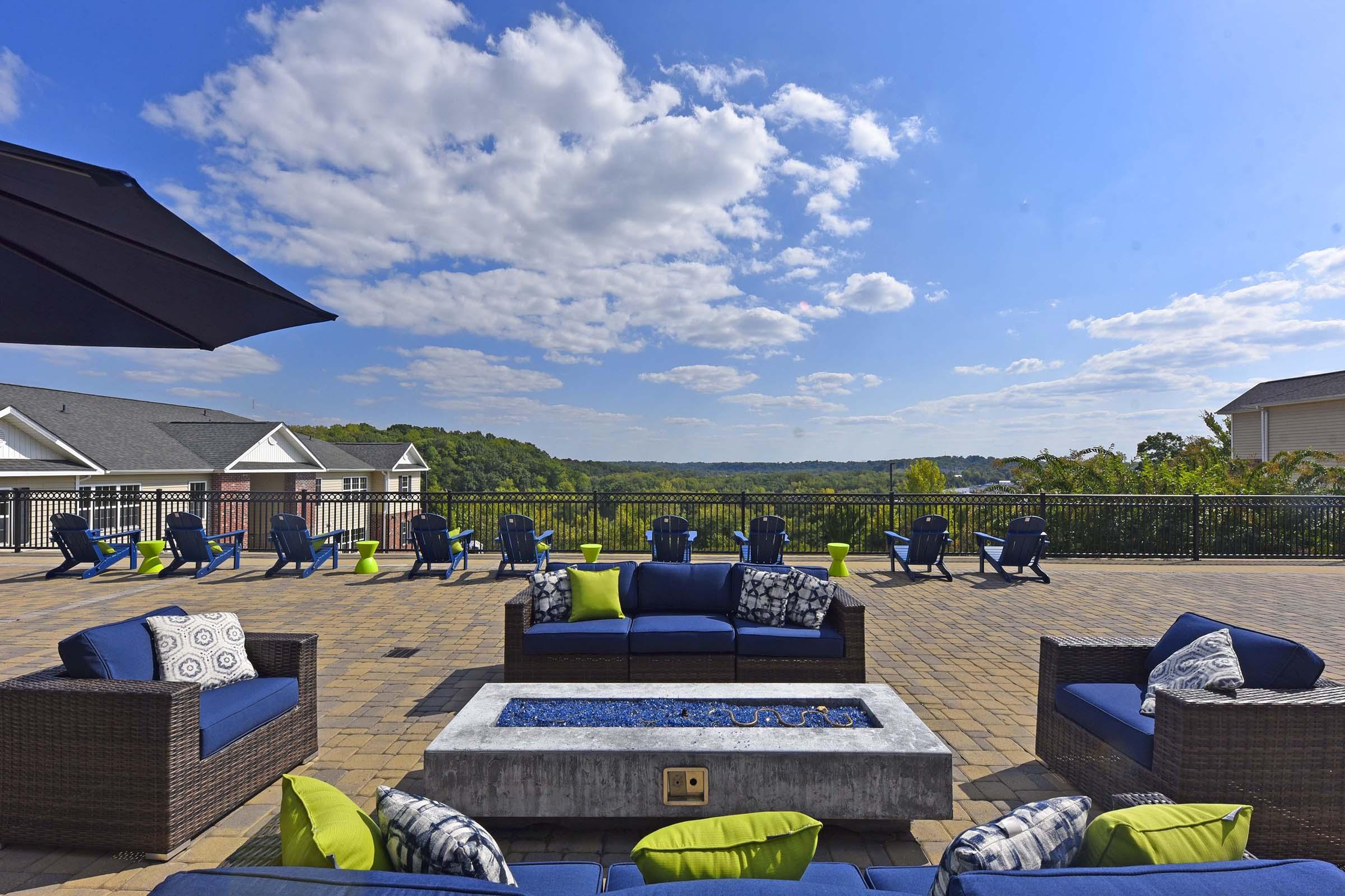 Outdoor lounge area with modern furniture, including blue and green cushions, surrounding a fire pit. In the background, there are several blue lounge chairs and a scenic view of greenery under a bright blue sky with fluffy white clouds.