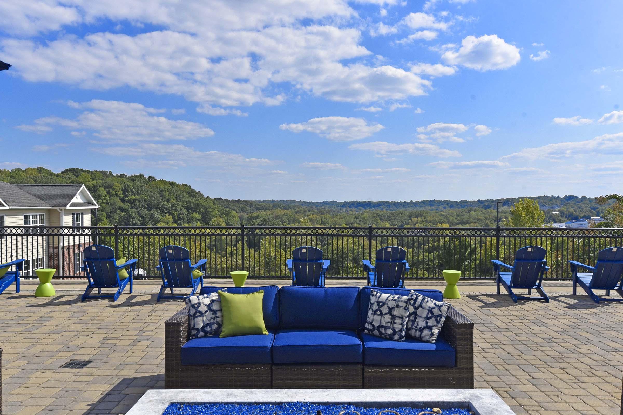 A scenic outdoor patio area with a blue couch featuring decorative pillows. Several blue Adirondack chairs and green side tables are arranged facing a panoramic view of trees and hills under a partly cloudy sky. The stone-paved floor adds to the inviting atmosphere.