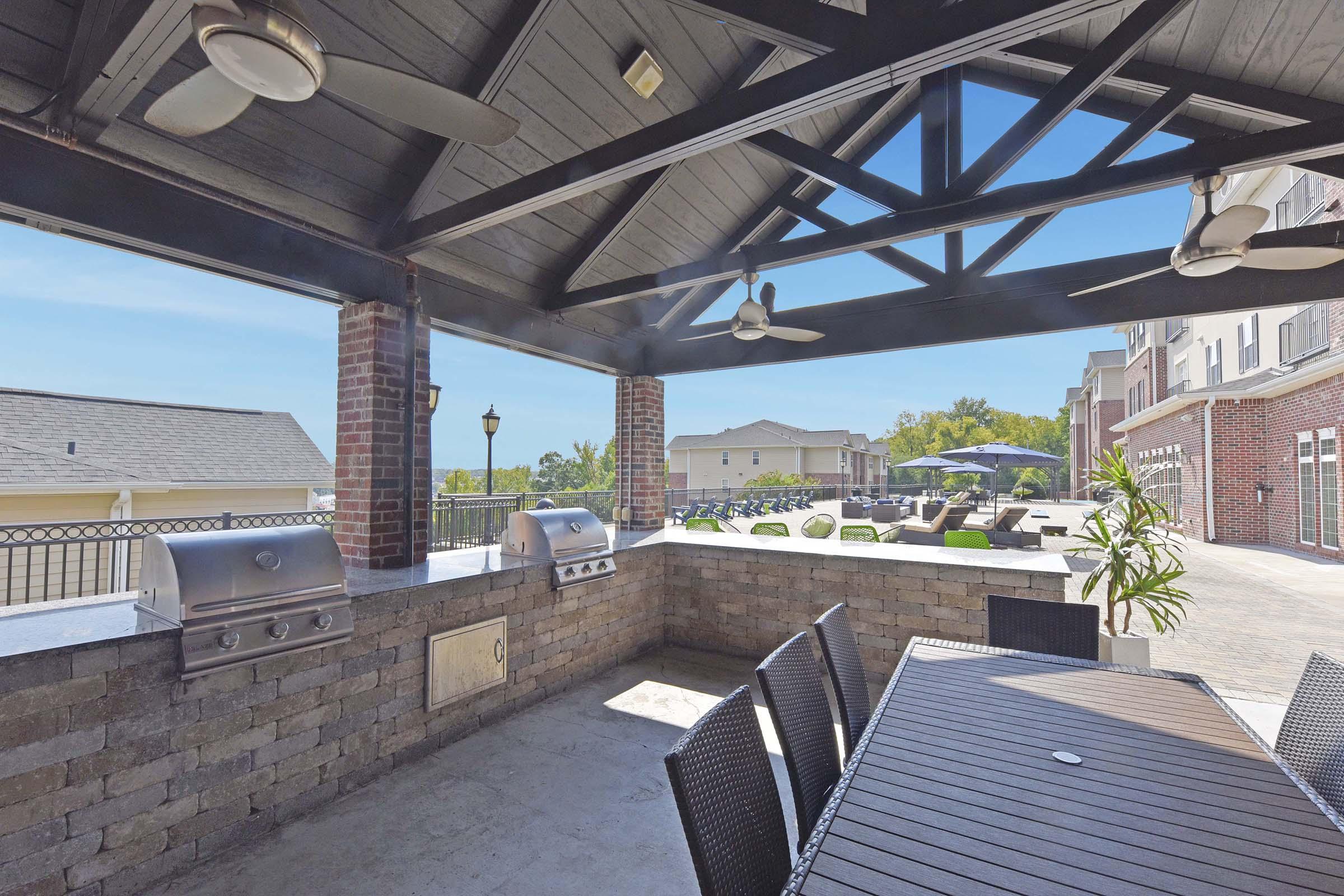 Outdoor grilling area featuring a large stone countertop with stainless steel barbecue grills, surrounded by black patio furniture. The space is shaded by a wooden pergola with ceiling fans, and the background showcases a well-maintained outdoor pool and lounge area under clear blue skies.