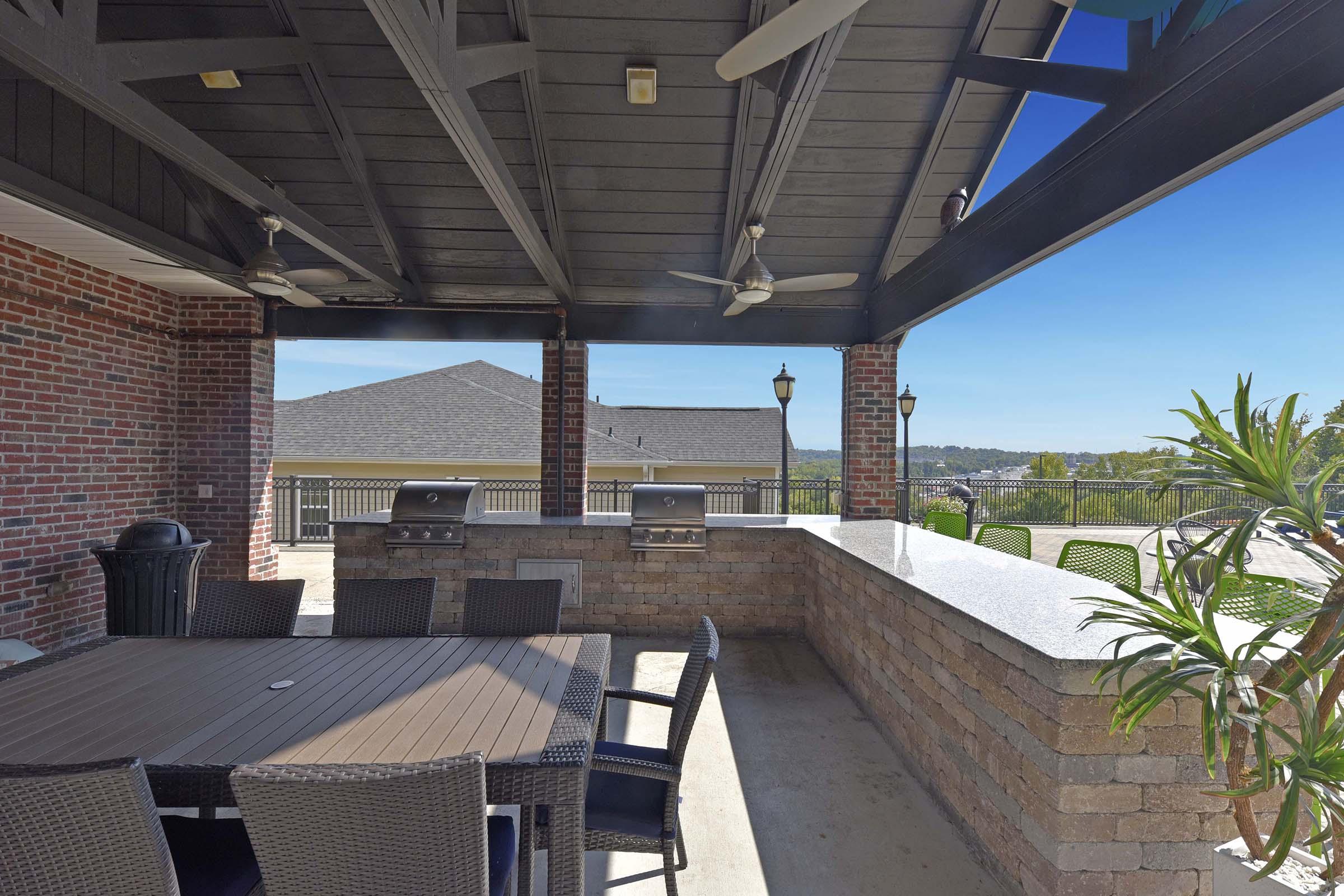 Outdoor patio area featuring a table with wicker chairs, a stone countertop, and built-in grills. The space is covered by a roof with exposed beams and has ceiling fans. In the background, there are hills and clear blue skies, providing a scenic view.