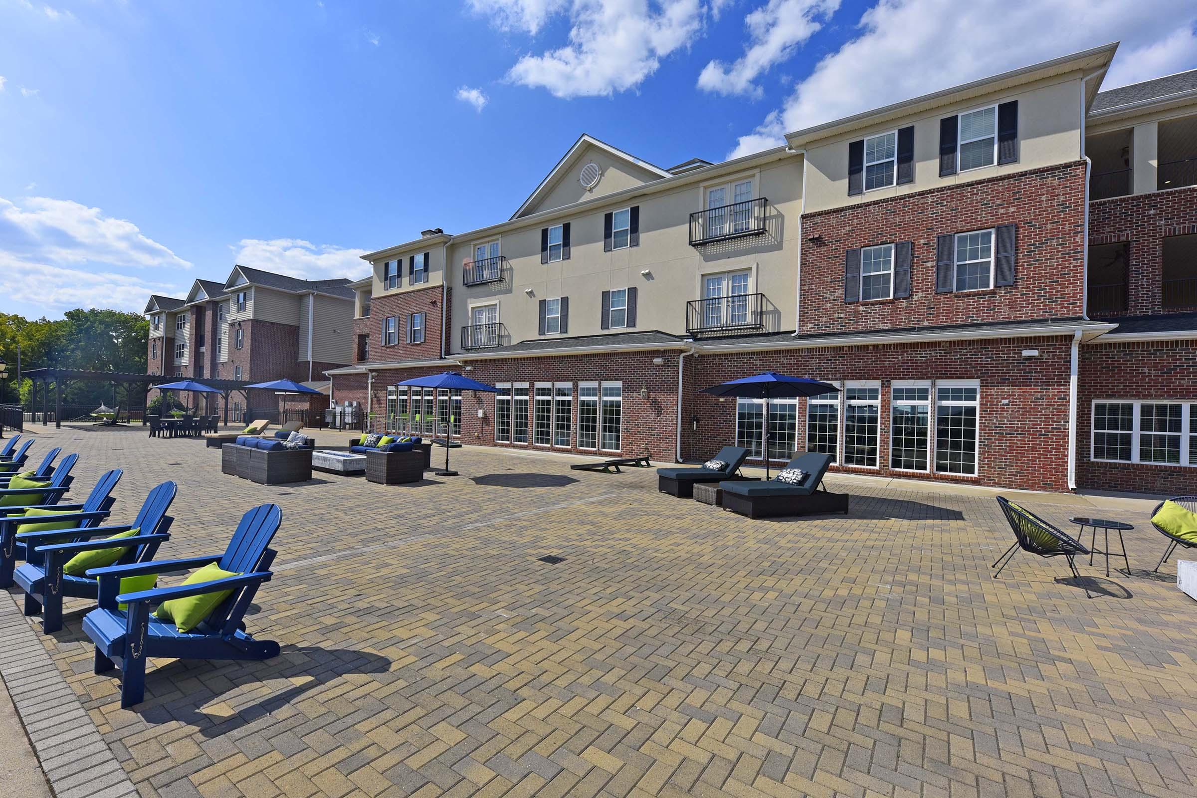 A brick building with multiple floors, featuring large windows and balconies. In the foreground, a paved area with lounge chairs and umbrellas, surrounded by greenery. The sky is clear with a few clouds, creating a sunny atmosphere.