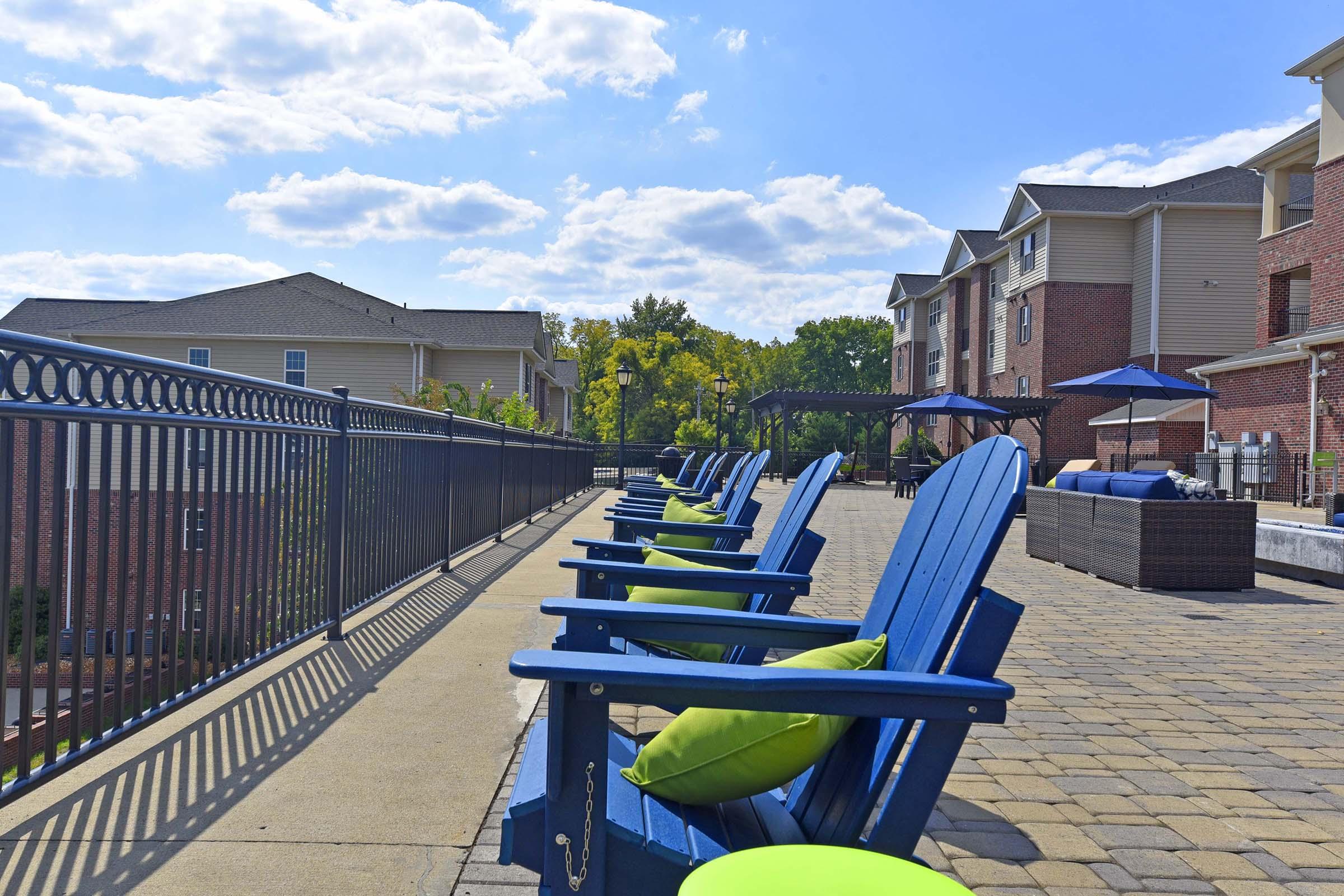 A sunny outdoor patio with blue lounge chairs featuring green cushions, overlooking a landscaped area. The setting includes a railing and nearby residential buildings under a partly cloudy sky, creating a relaxing and inviting atmosphere.