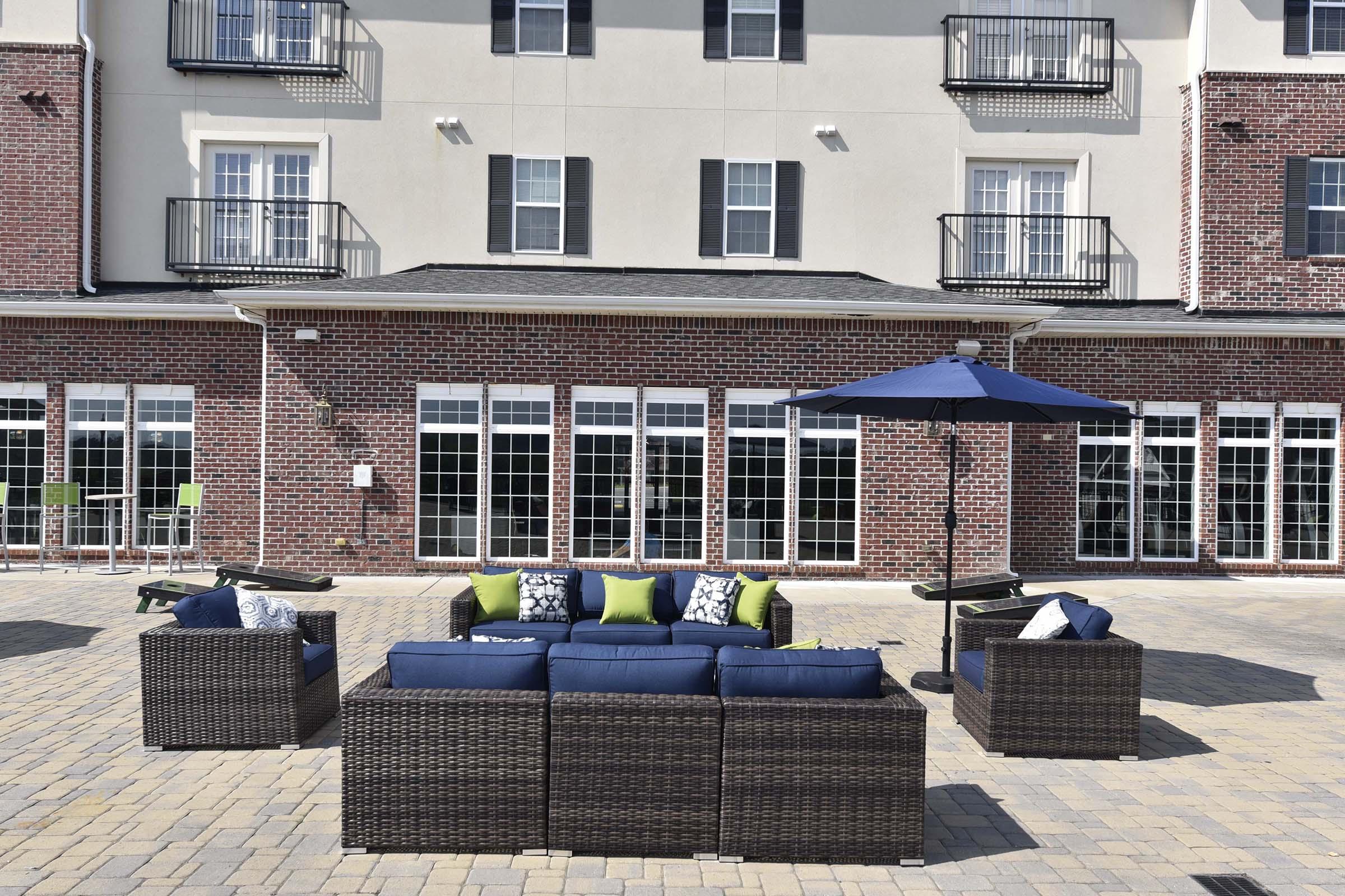 A stylish outdoor seating area featuring a sectional sofa with navy cushions and green accent pillows, surrounded by a brick building. An umbrella shades the seating, and large windows with black shutters reflect the sunlight. Paved stone flooring enhances the modern and inviting atmosphere.