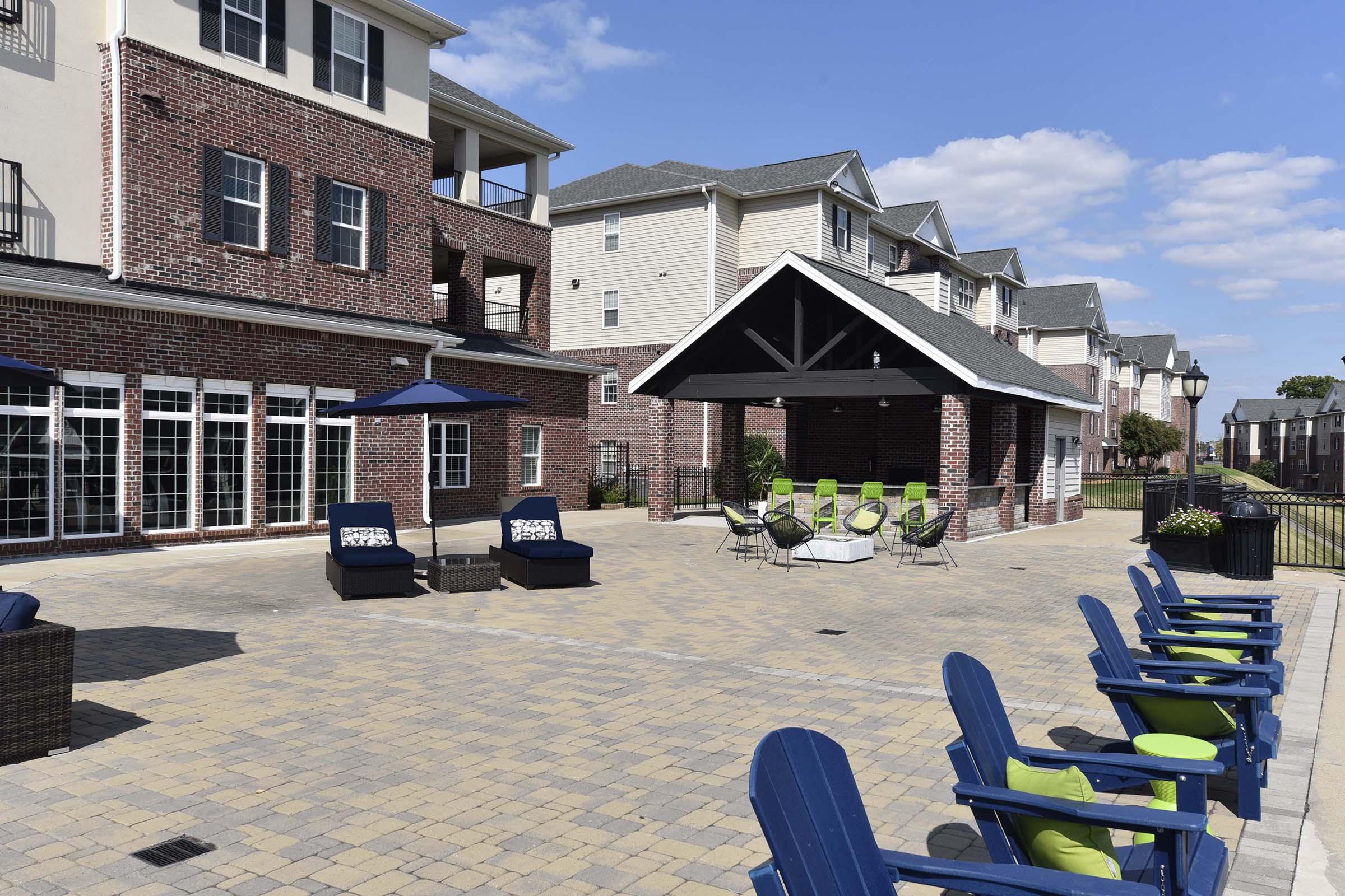 A spacious outdoor patio area featuring blue lounge chairs and green accents, surrounded by brick buildings. The scene includes a covered pavilion with seating and tables, set against a clear blue sky and well-maintained landscaping.