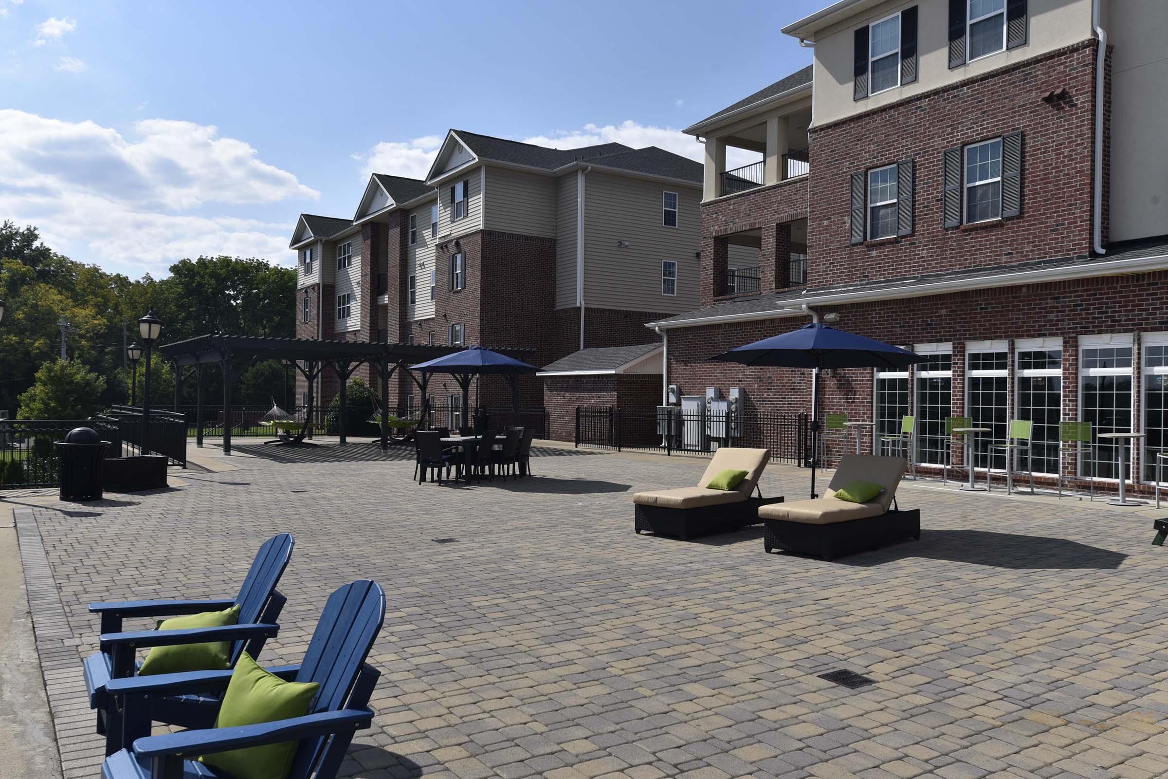 A sunny outdoor lounge area featuring blue adirondack chairs, two lounge chairs with green pillows, and a brick patio. There are patio tables and umbrellas, surrounded by a landscaped area with trees, and a multi-story building in the background. Clear skies and pleasant weather create a relaxing atmosphere.